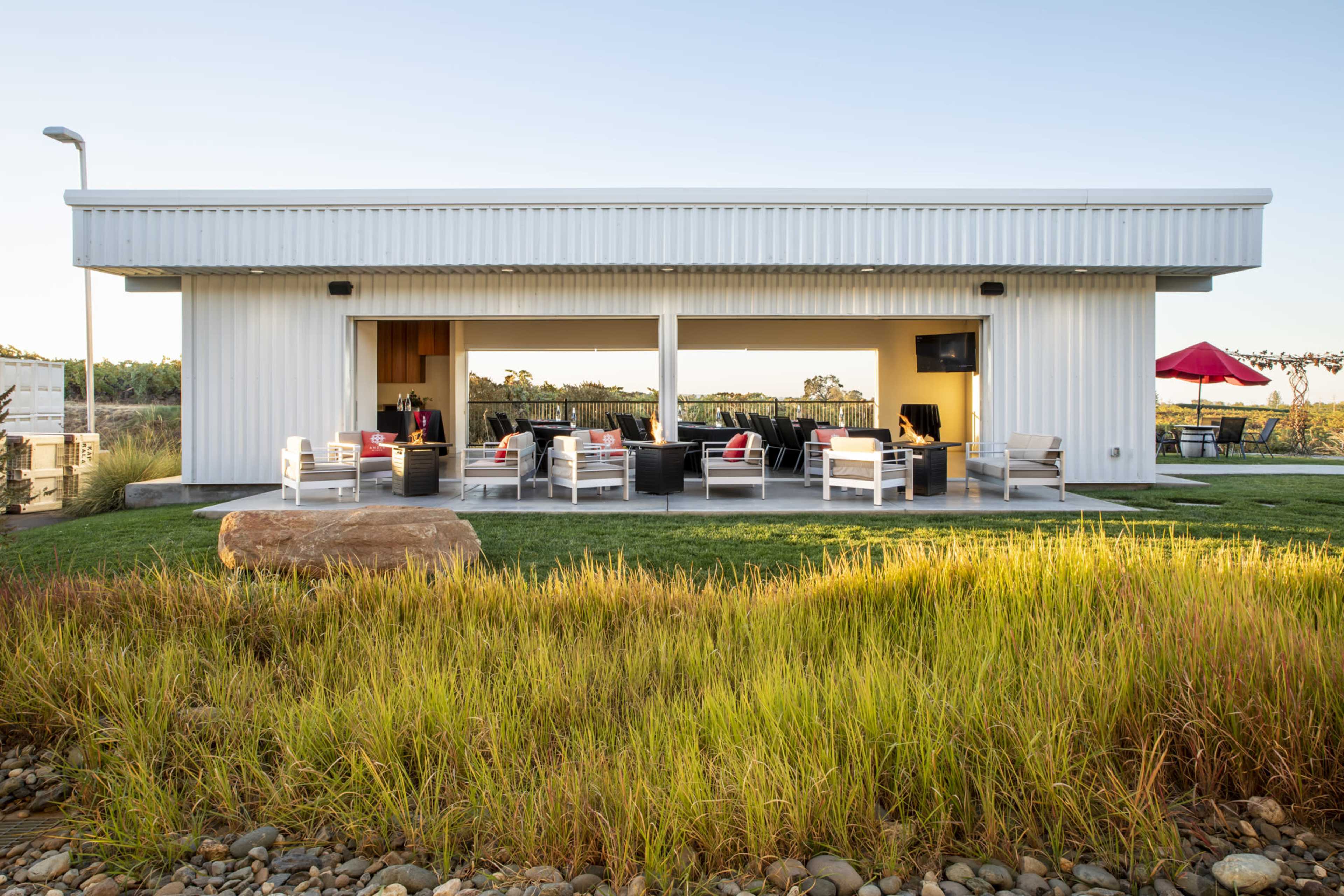 A modern, single-story structure with large openings and outdoor seating overlooks a grassy area and is set against a clear sky.