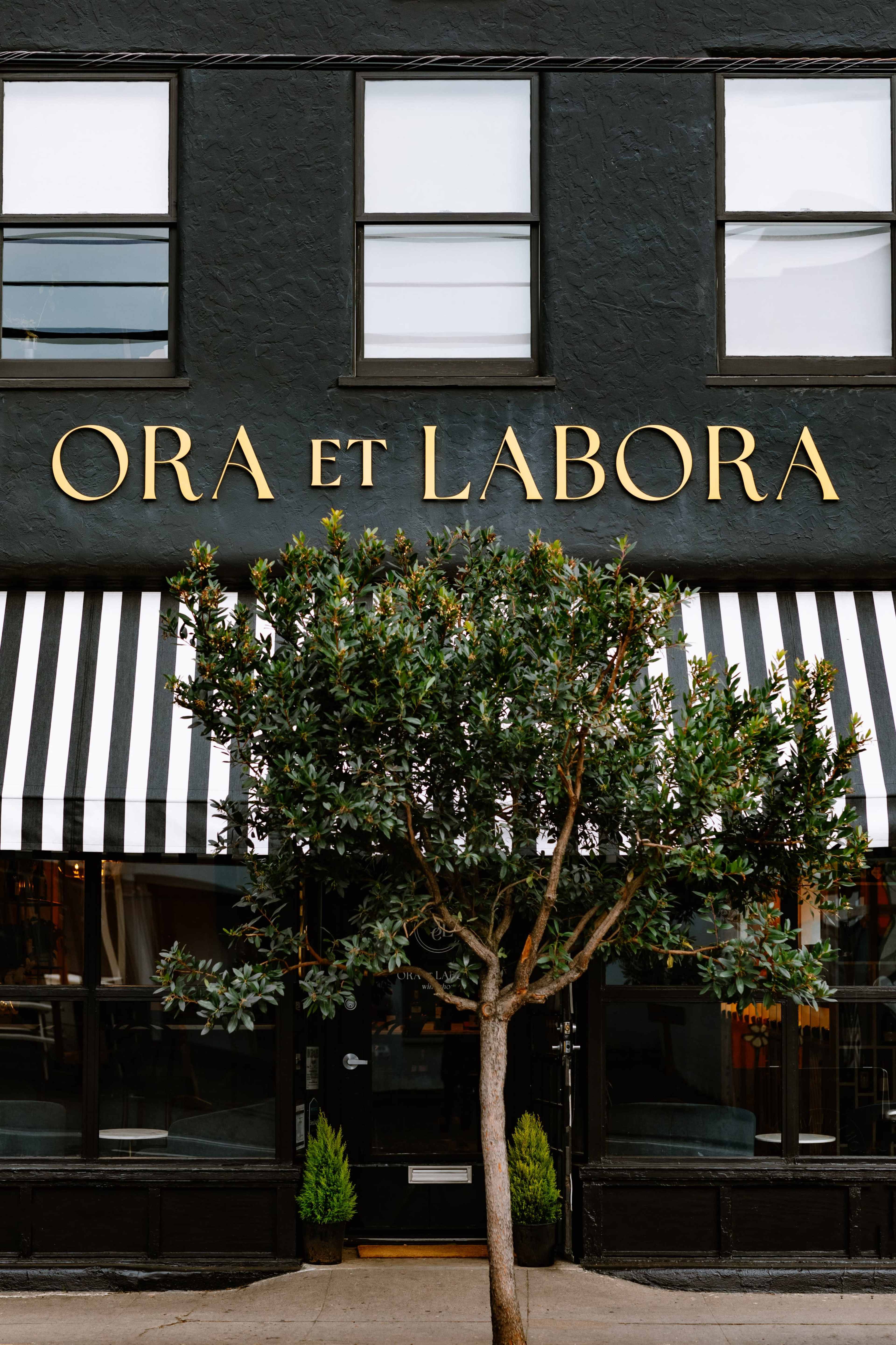 A building front with the inscription "ORA ET LABORA" and striped awnings, accompanied by a small tree in front.