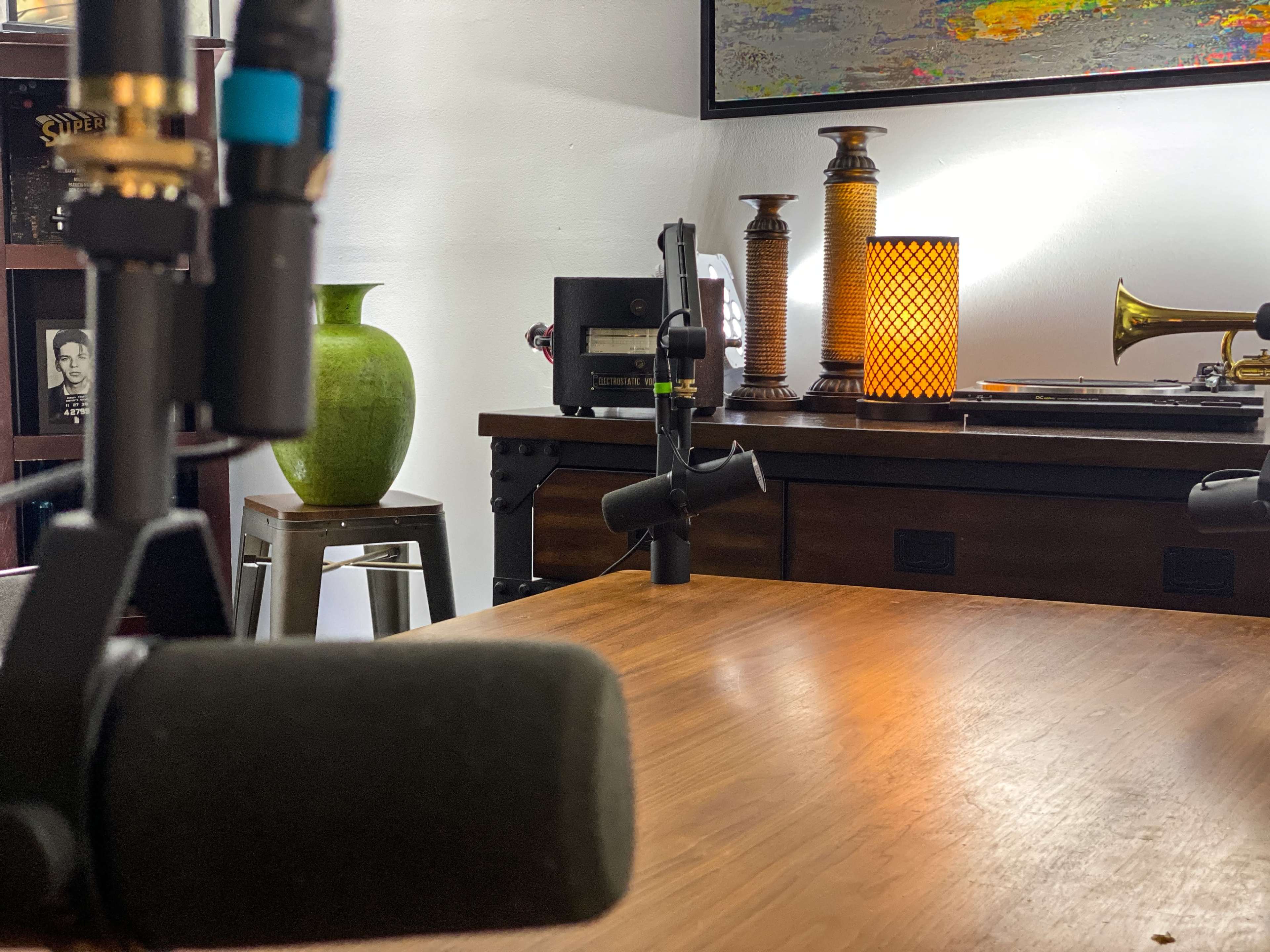 A wooden table with microphones in the foreground and various decorative items, including a green vase and vintage lamps, in the background.