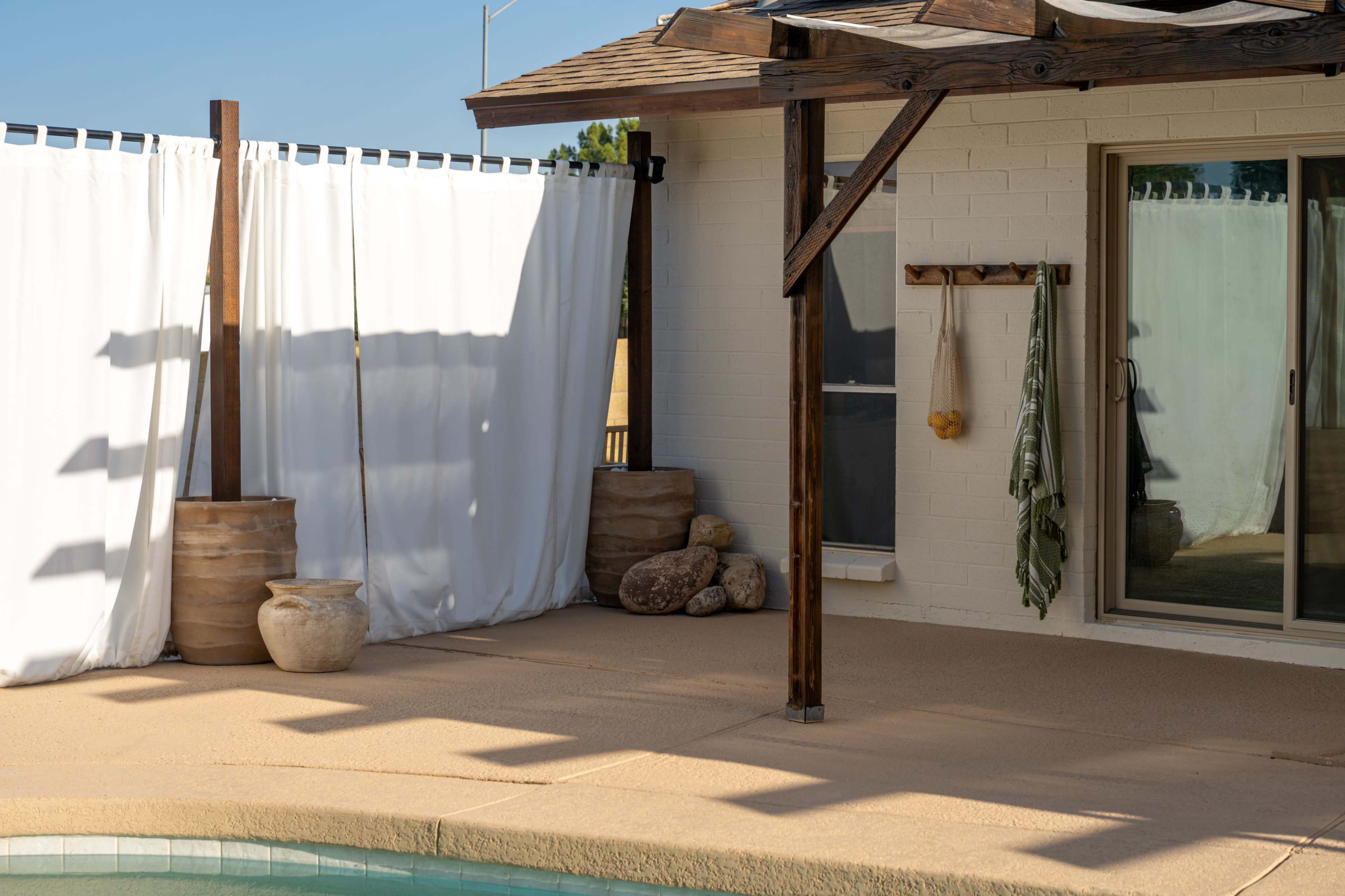 The scene features a partially shaded patio area with white curtains, decorative pots, and a view of a sliding glass door.