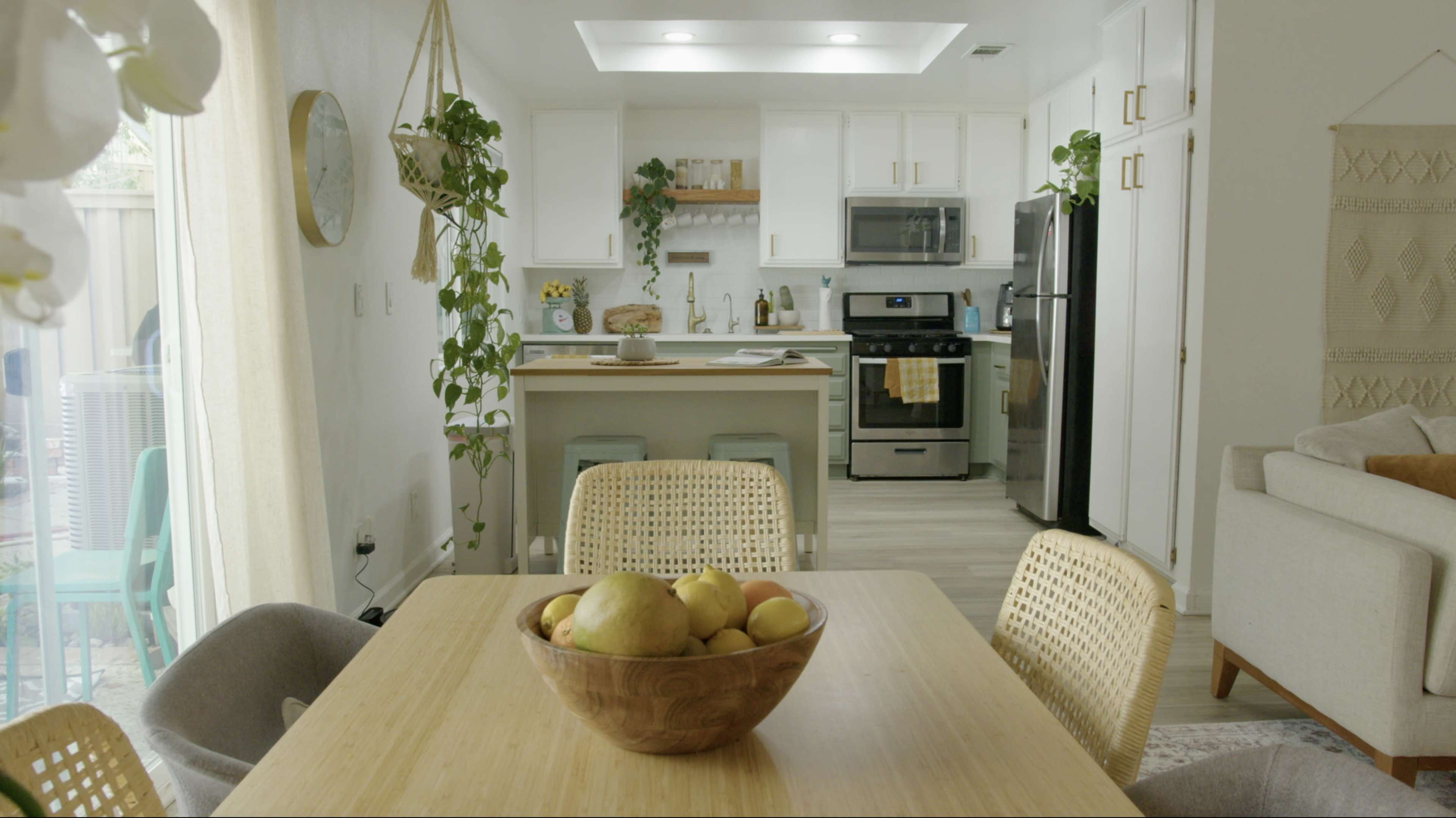 A kitchen and dining area feature a wooden table with a fruit bowl, surrounded by wicker chairs, while the kitchen has white cabinets and stainless steel appliances.