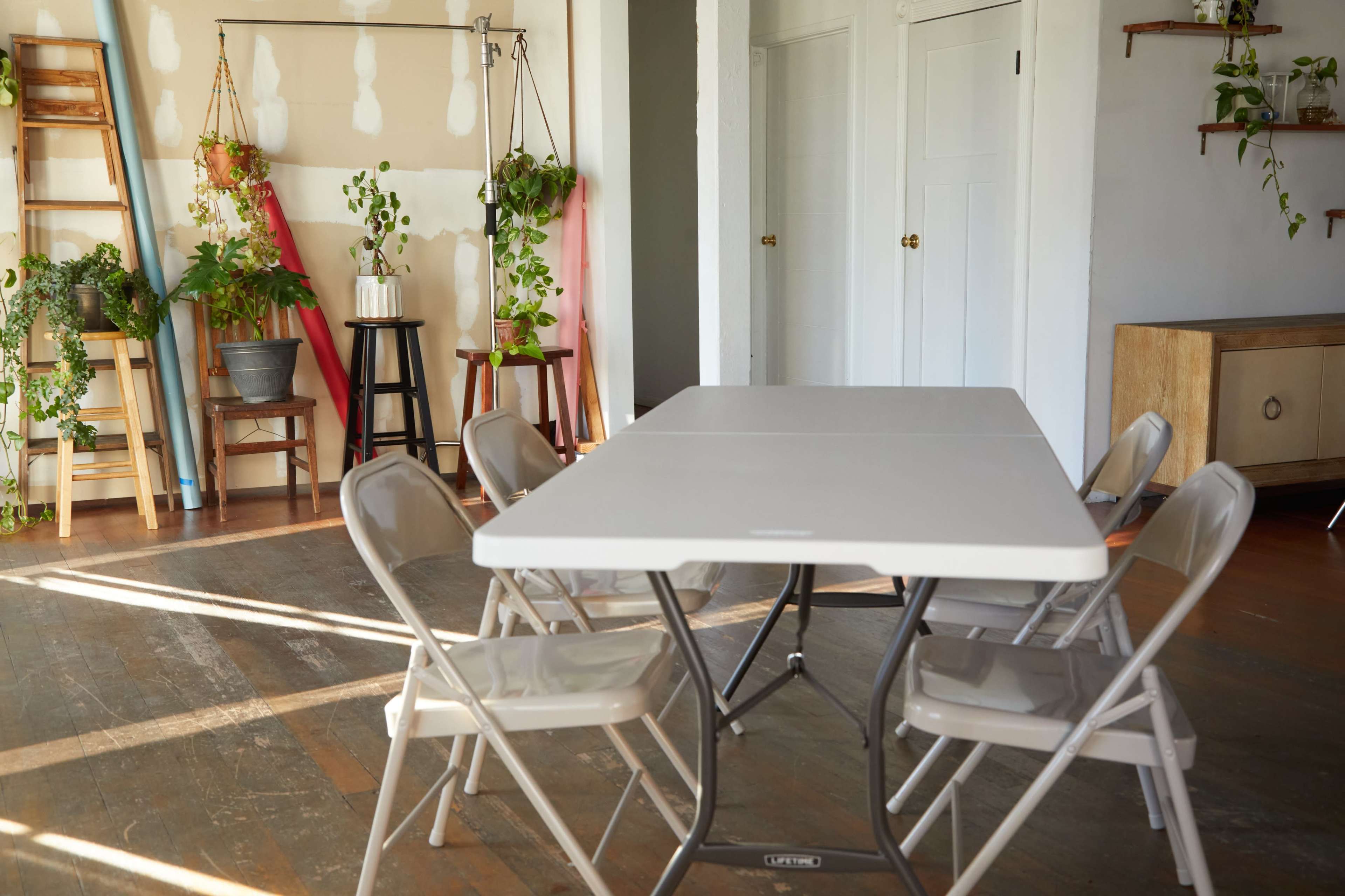 A white folding table is set up in a room with several metal folding chairs and various plants displayed on shelves and stands.