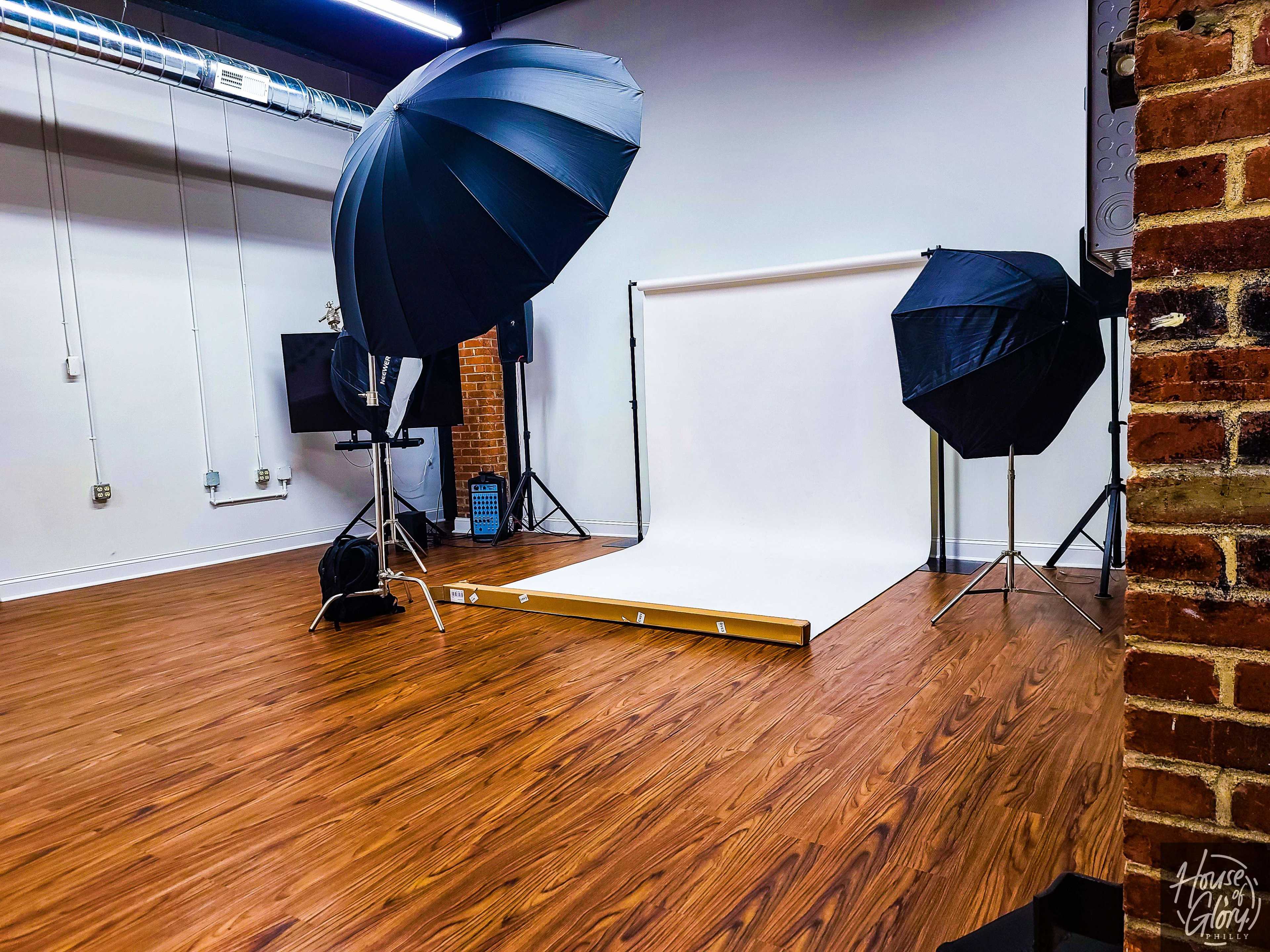 A photography studio features a white backdrop, two large black umbrellas, and wooden flooring.