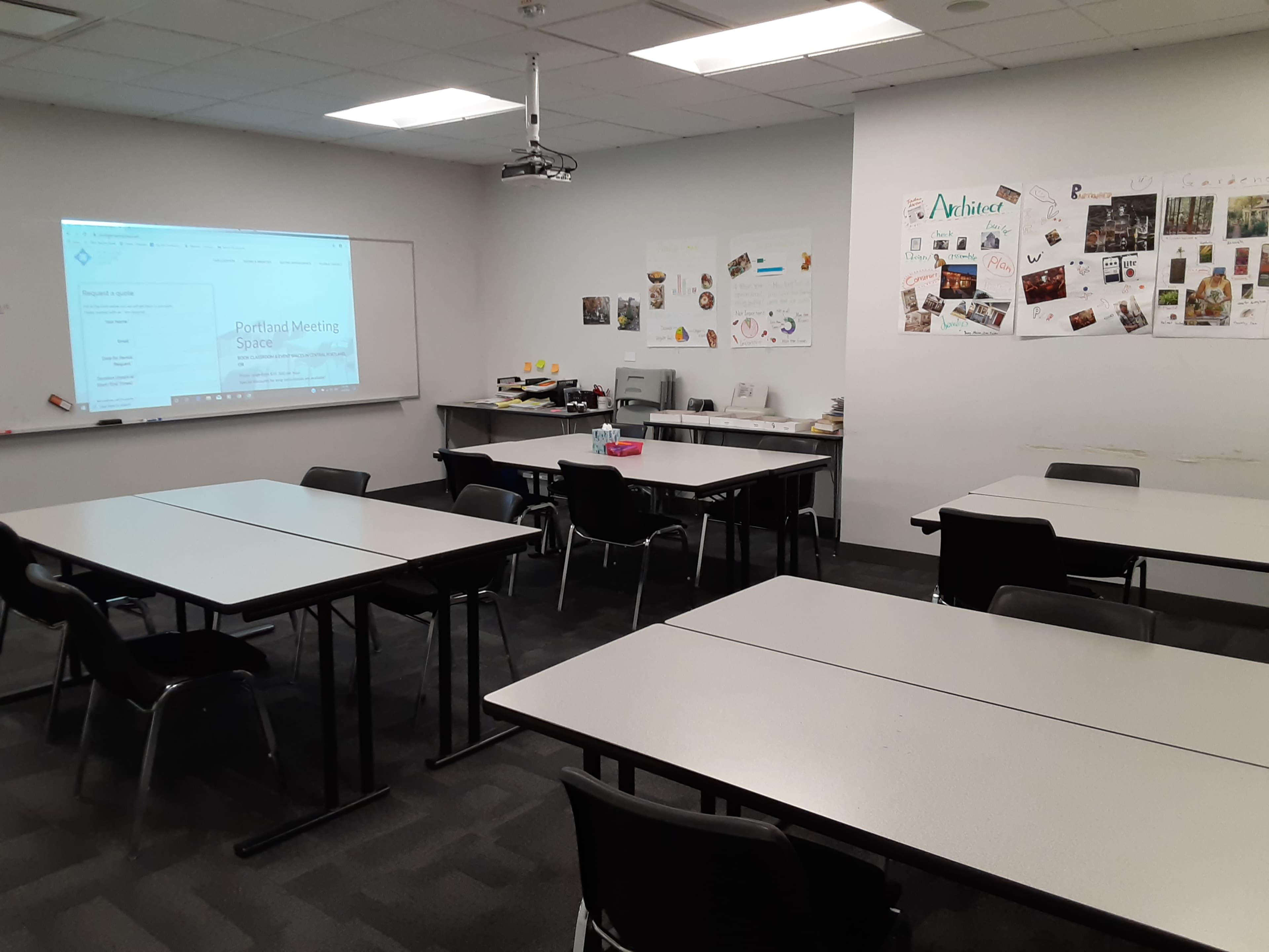 A classroom setting features multiple tables arranged with chairs, a projector screen displaying content, and a wall adorned with various images and decorations.