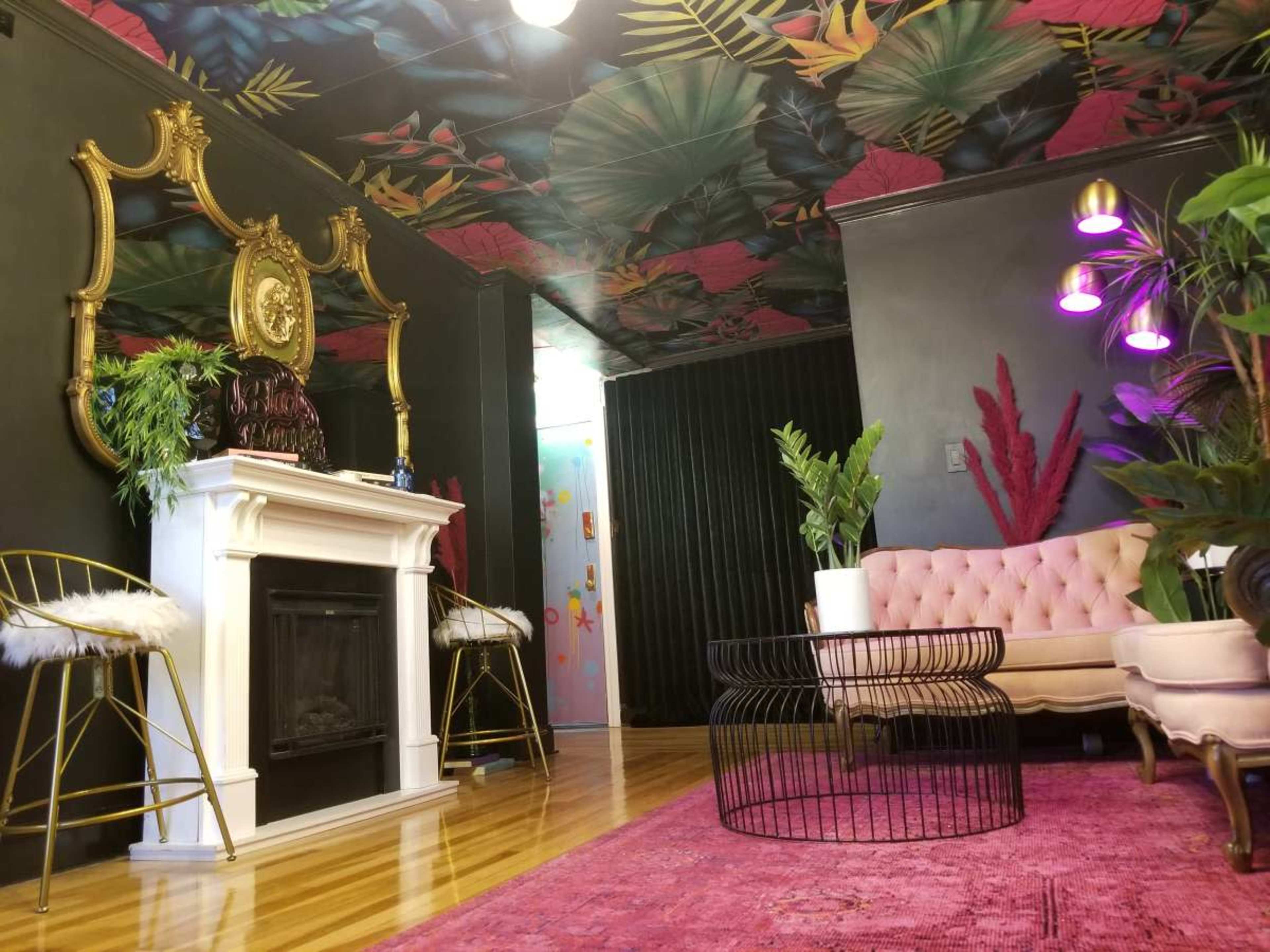 The image shows a stylish room featuring a pink tufted couch, a modern black coffee table, a decorative fireplace, and vibrant plants against dark walls and a colorful ceiling.