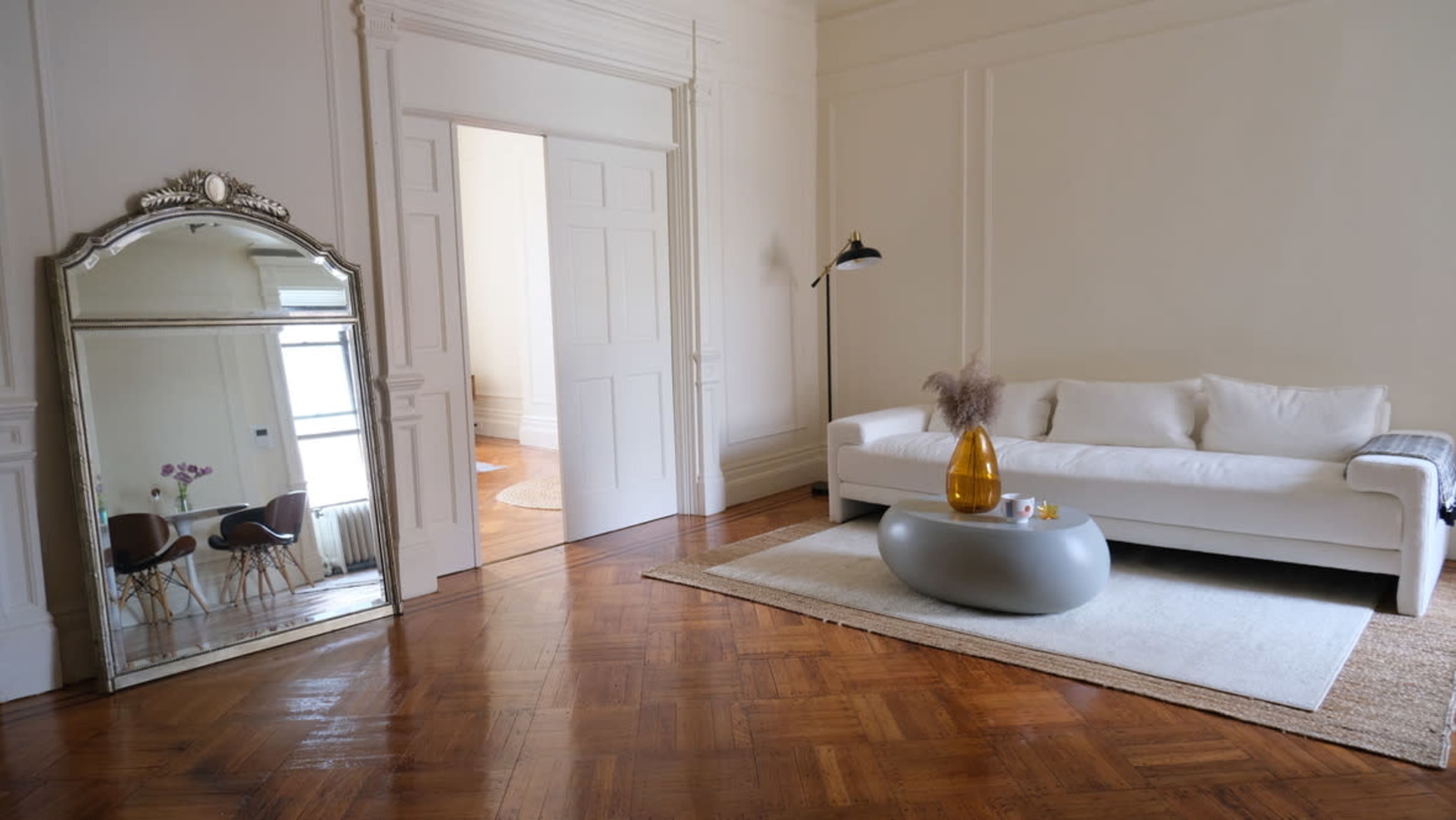 The room features a white couch, a round coffee table with a vase, and a large mirror against a wall, complemented by wooden flooring.