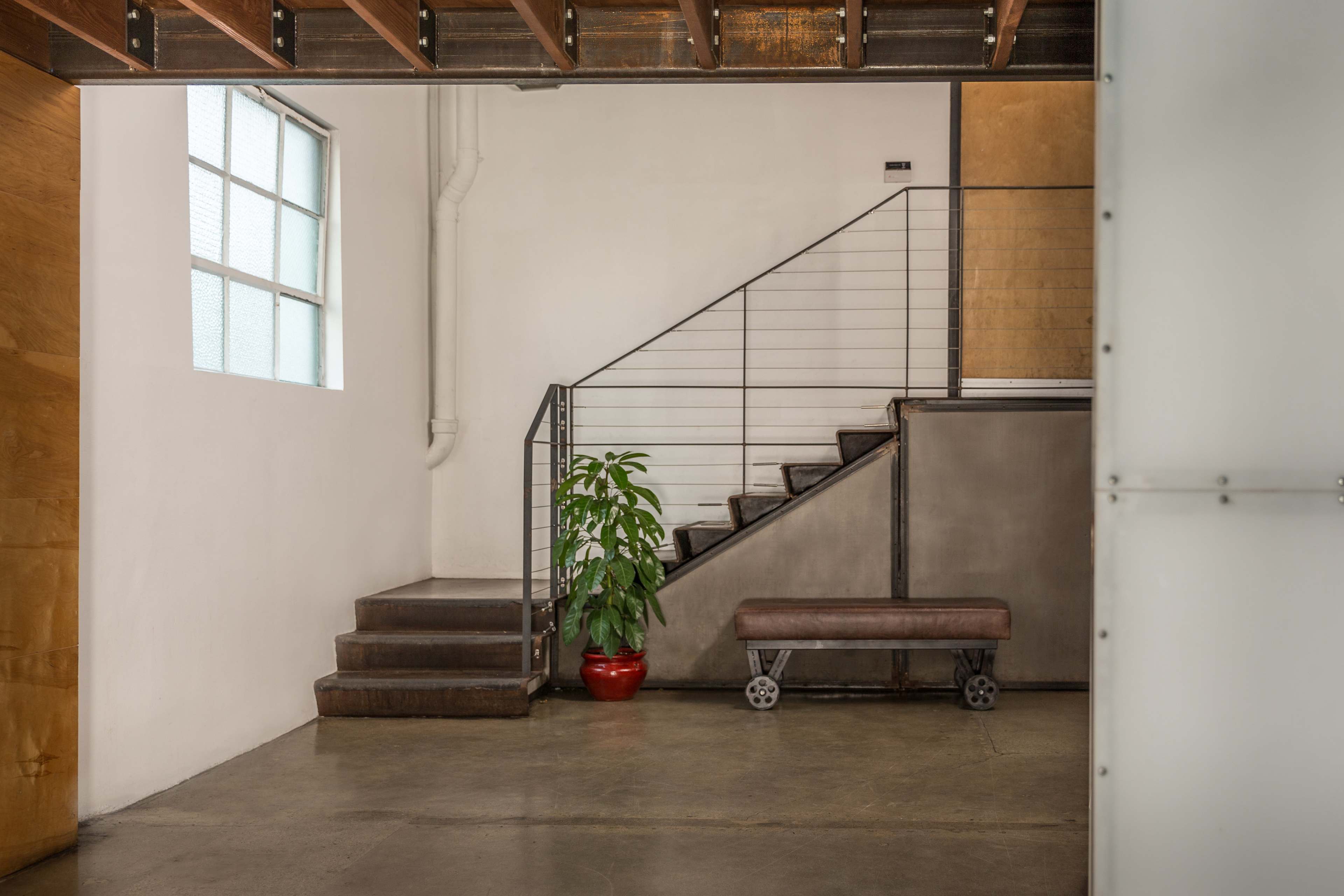 A staircase with a metal railing is positioned next to a potted plant in a room with exposed beams and a large window.
