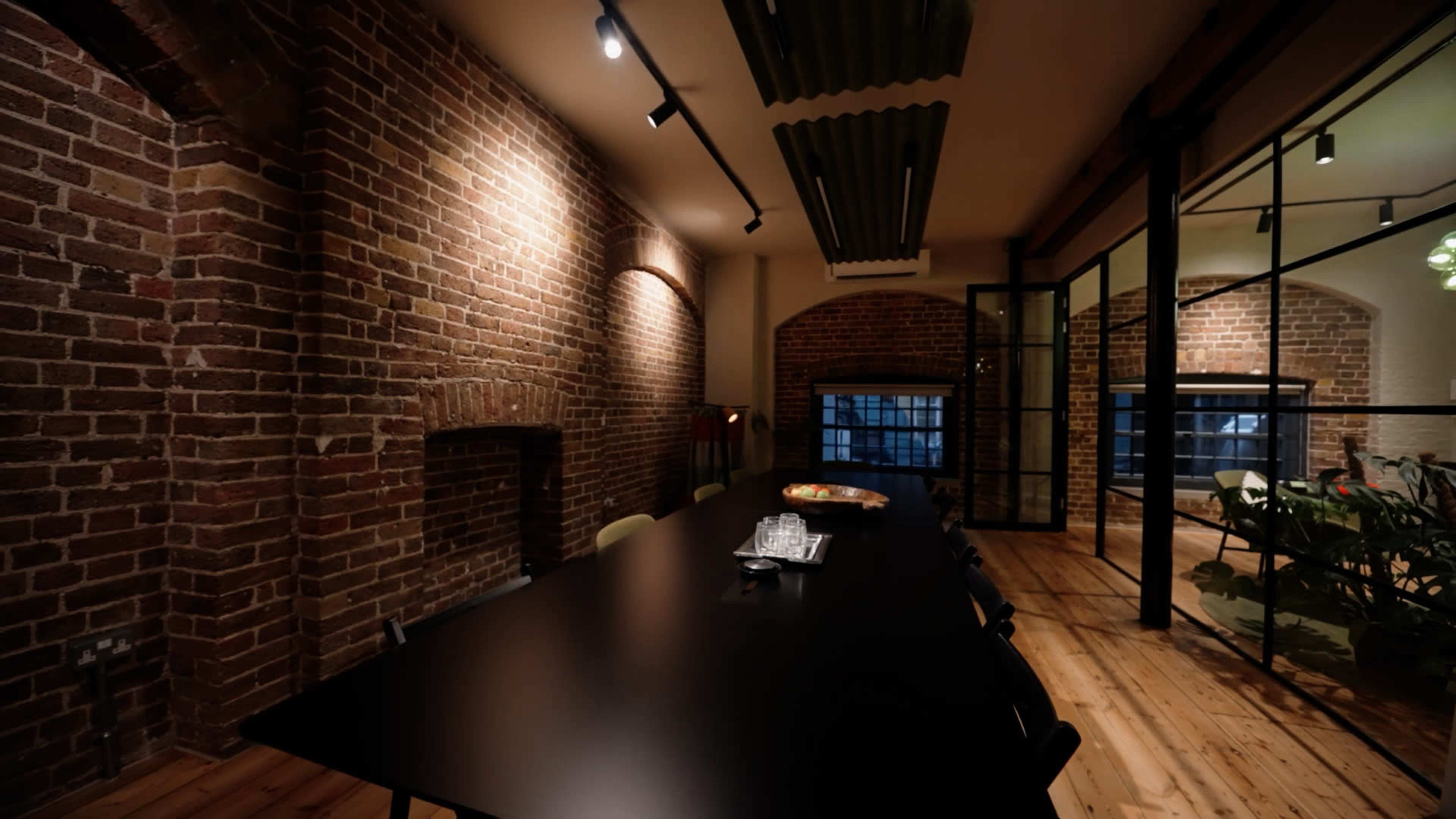 The image shows a modern dining area with a long black table, exposed brick walls, and wooden flooring, illuminated by track lighting.
