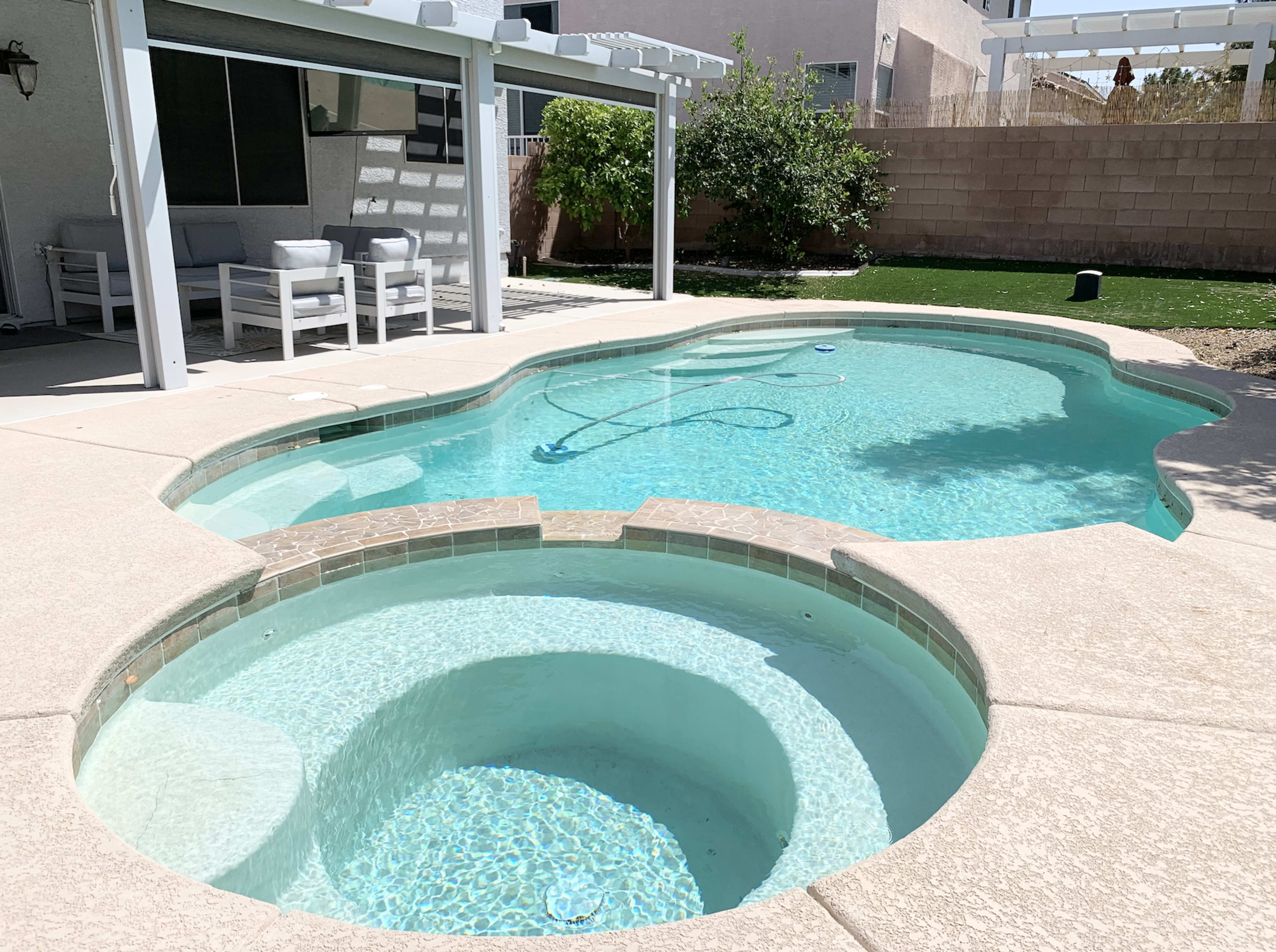 A backyard swimming pool with a connected spa, surrounded by a concrete patio and a shaded seating area.