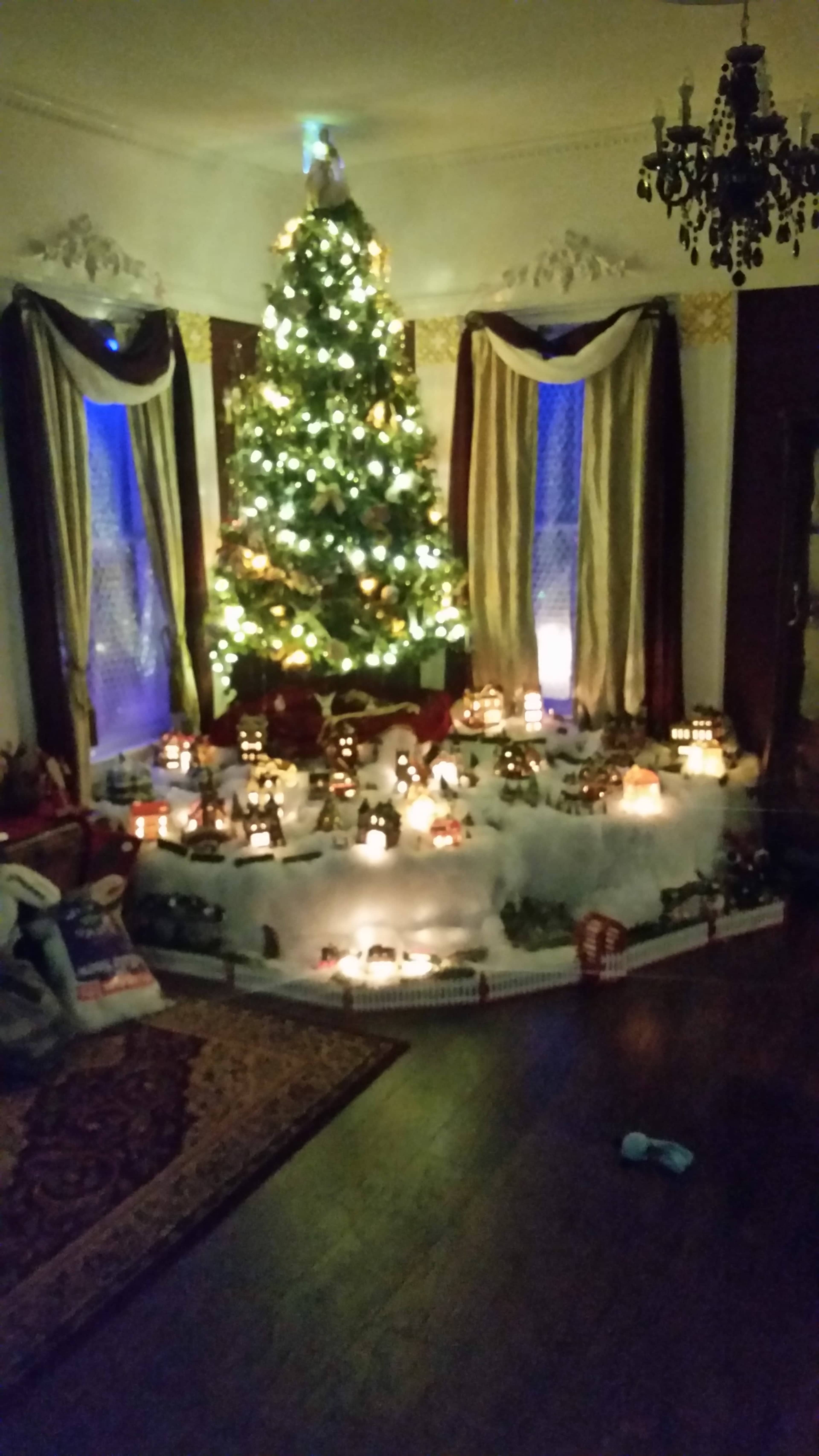 A decorative living room features a Christmas tree adorned with lights, surrounded by a snowy village scene with miniature buildings and figures.