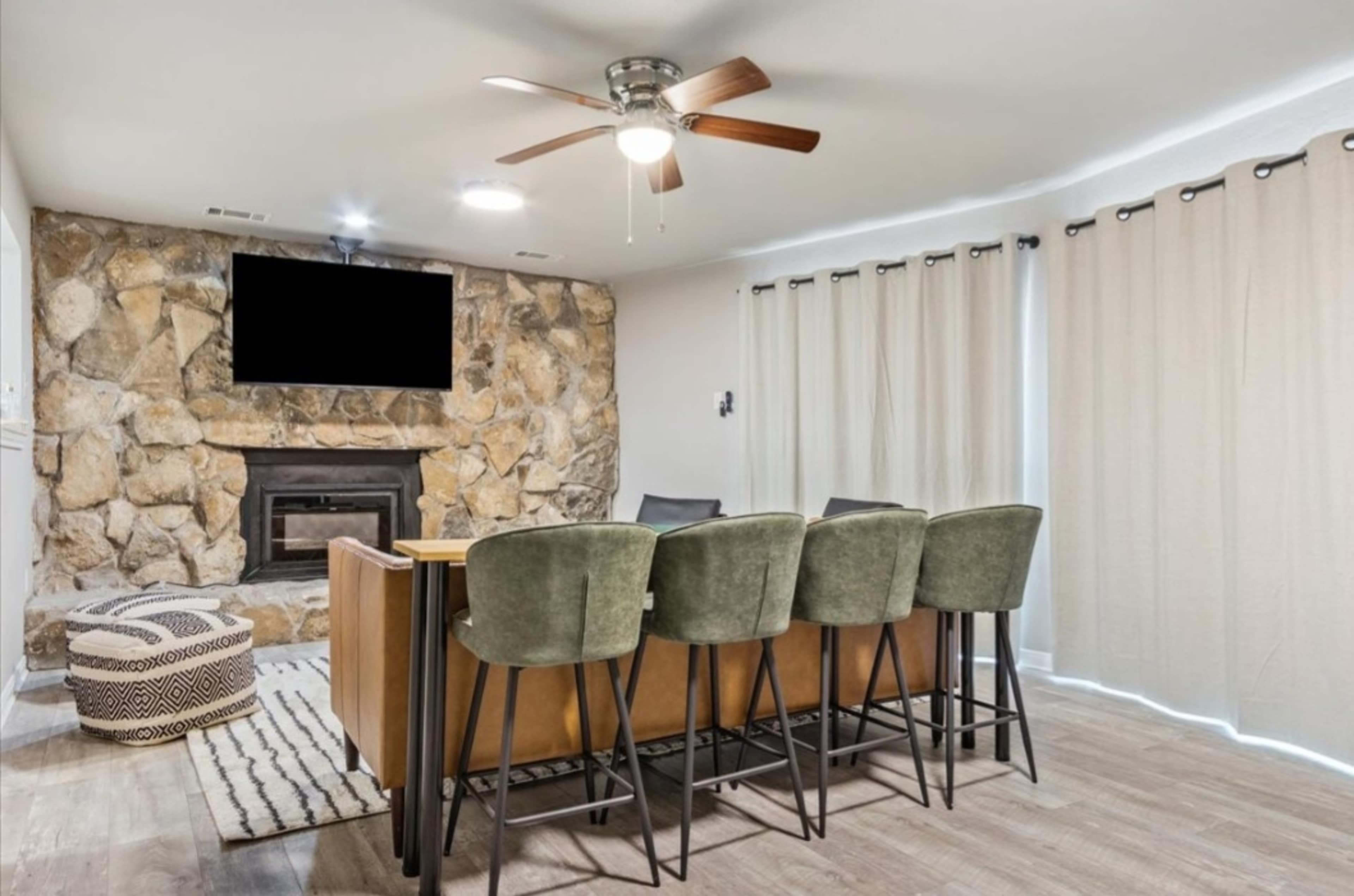 A living room with a stone fireplace, a television mounted above it, a dining table with high chairs, and beige curtains covering the windows.