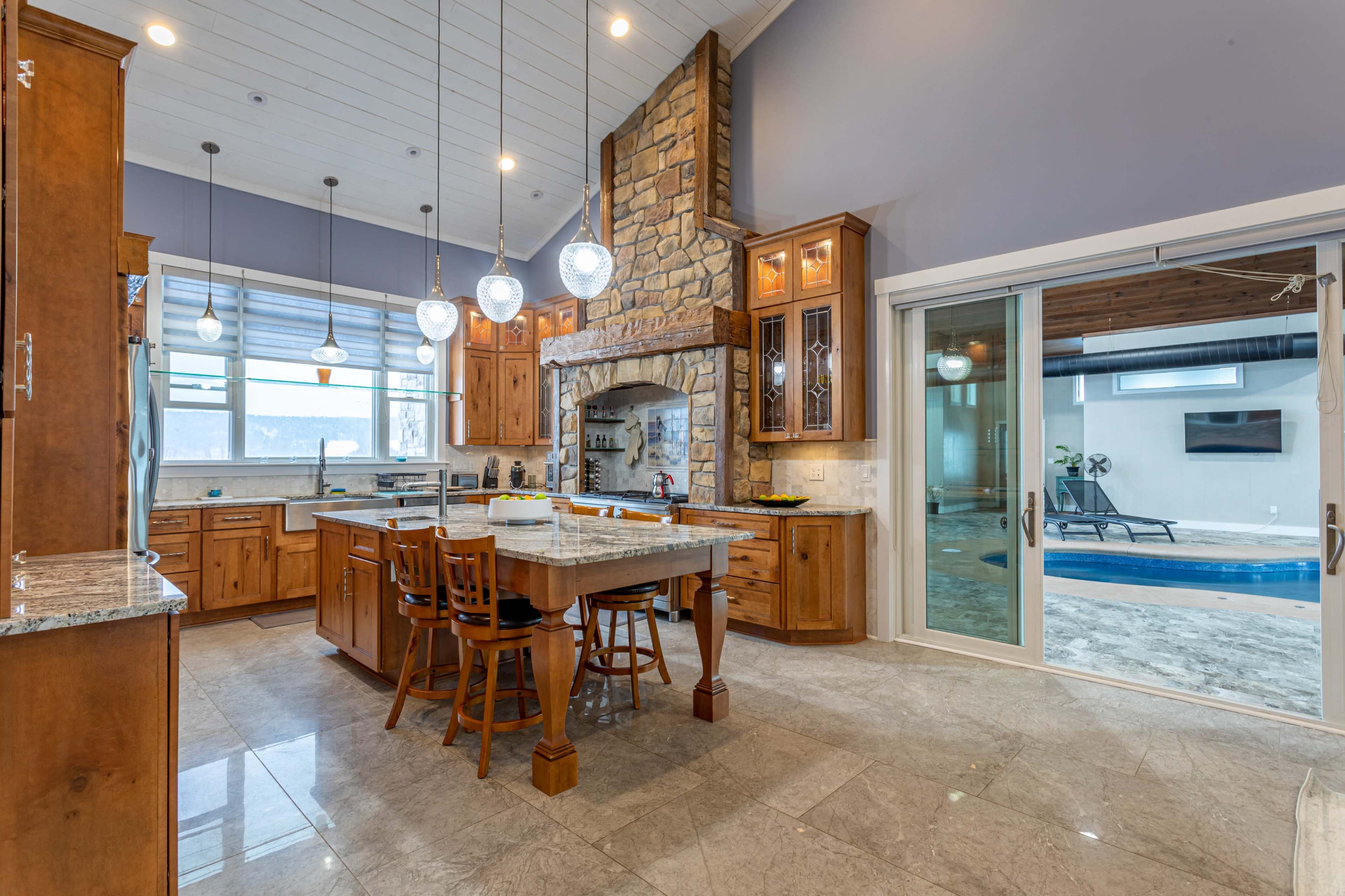 A modern kitchen features wooden cabinets, a large central island with seating, and a stone fireplace, leading to a sliding glass door that opens to a pool area.