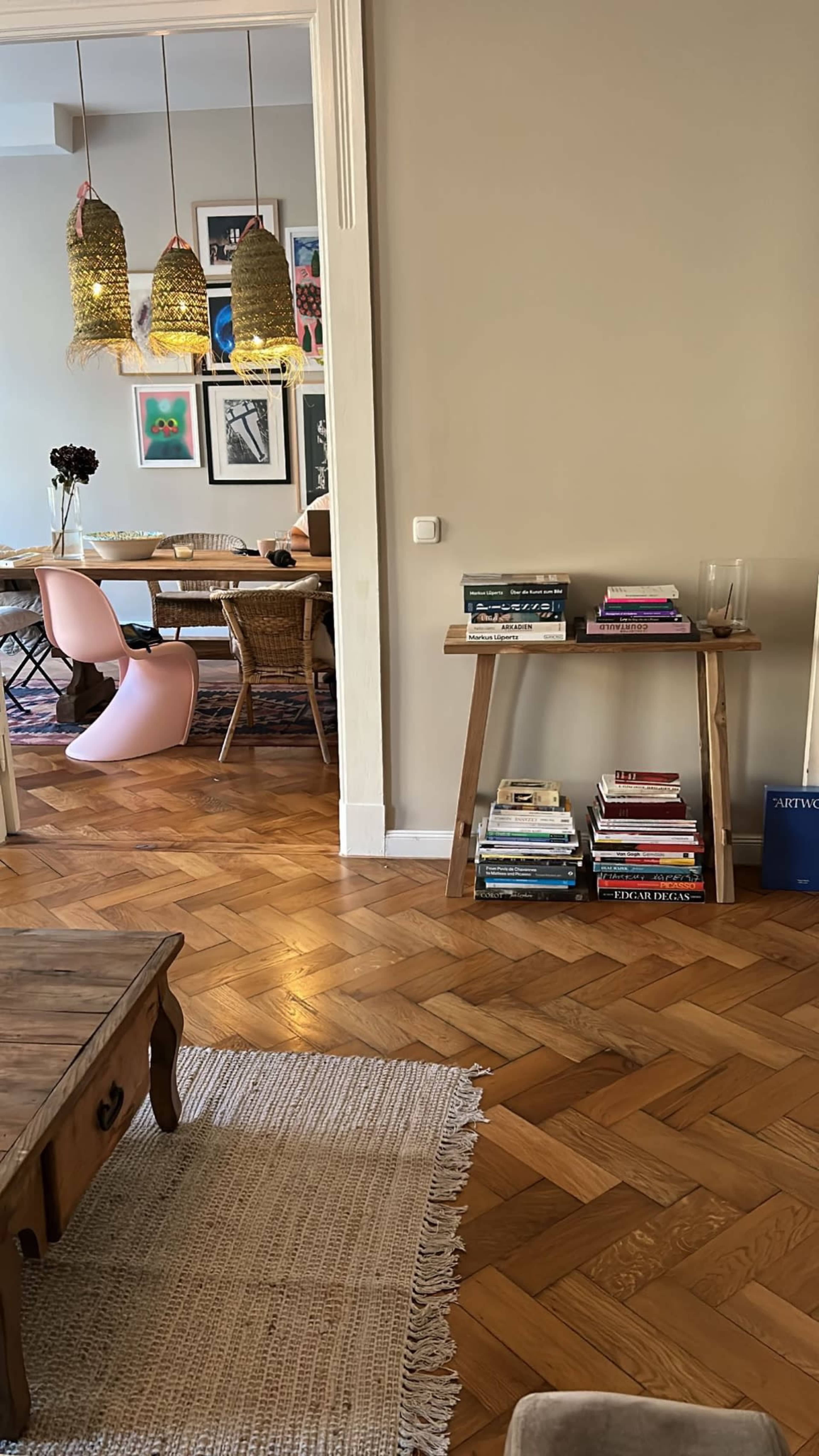 A living room features a wooden coffee table and a bookshelf, with a dining area visible in the background, adorned with decorative pendant lights and artwork on the walls.