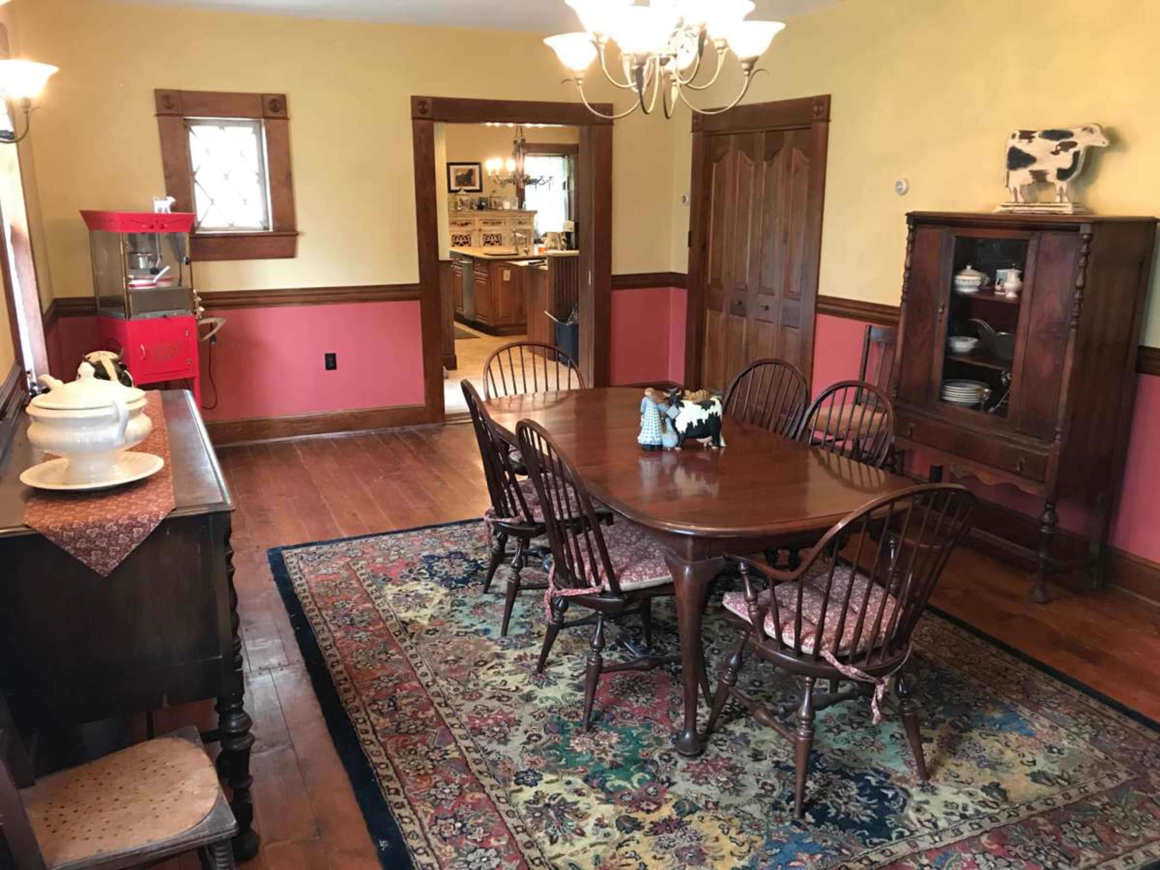 The image shows a dining room with a wooden table surrounded by six chairs, a sideboard, and a decorative rug on the hardwood floor, with yellow and red walls.