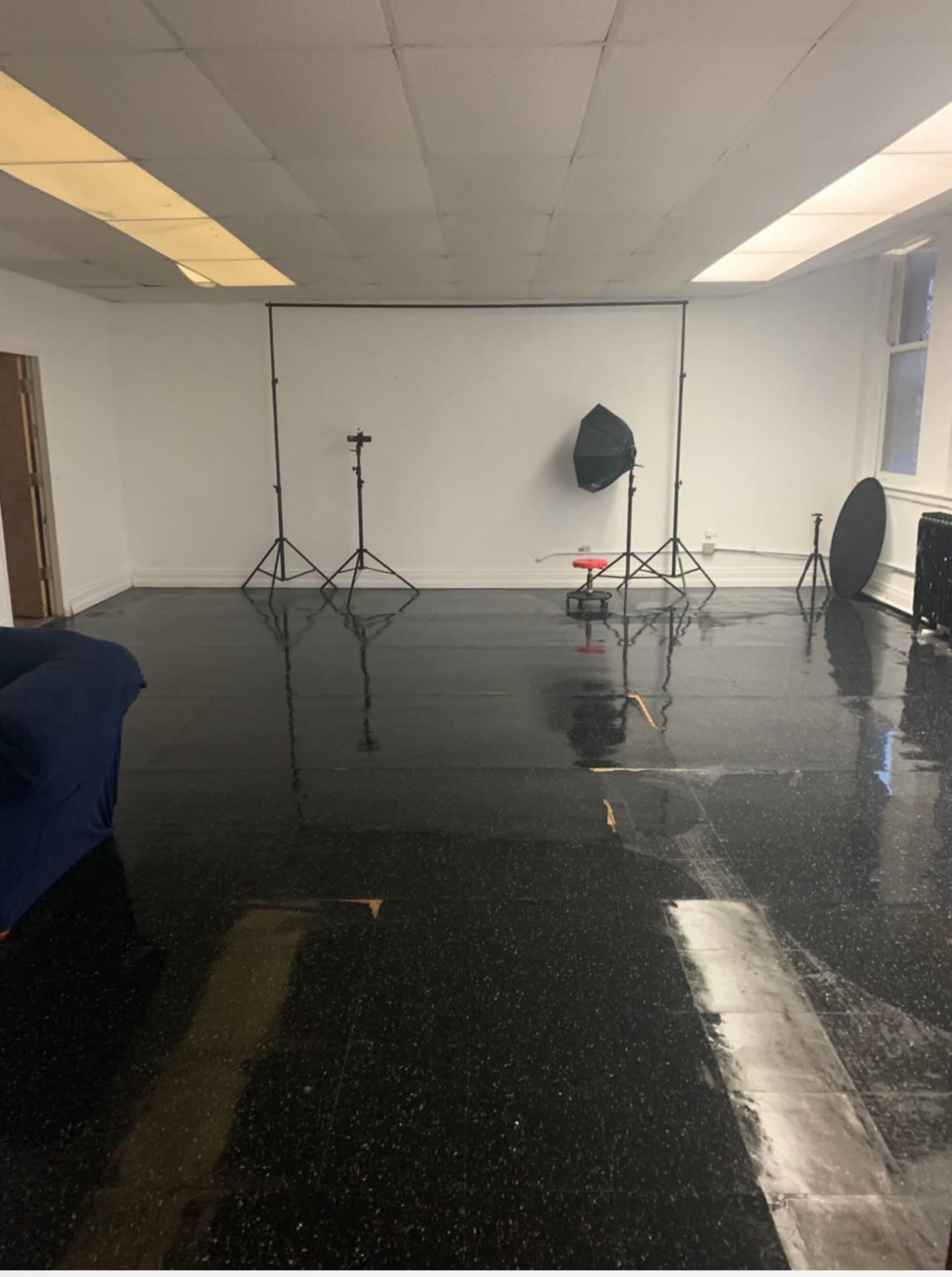 A photography studio with a glossy black floor, backdrop stands, and lighting equipment set up against a plain white wall.
