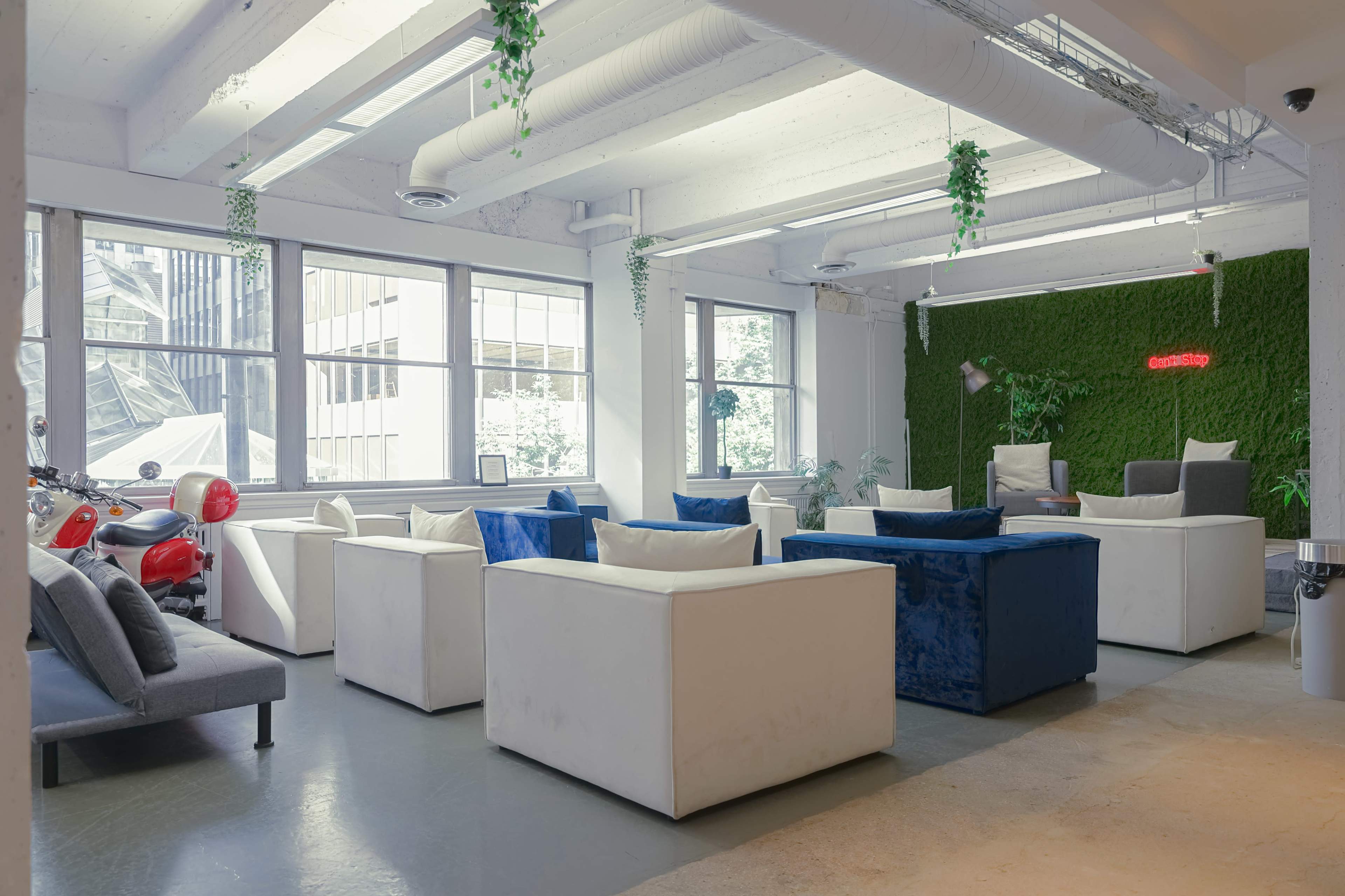 The image shows a modern lounge area with several white and blue upholstered seating arrangements, large windows, and a green wall with decorative plants.