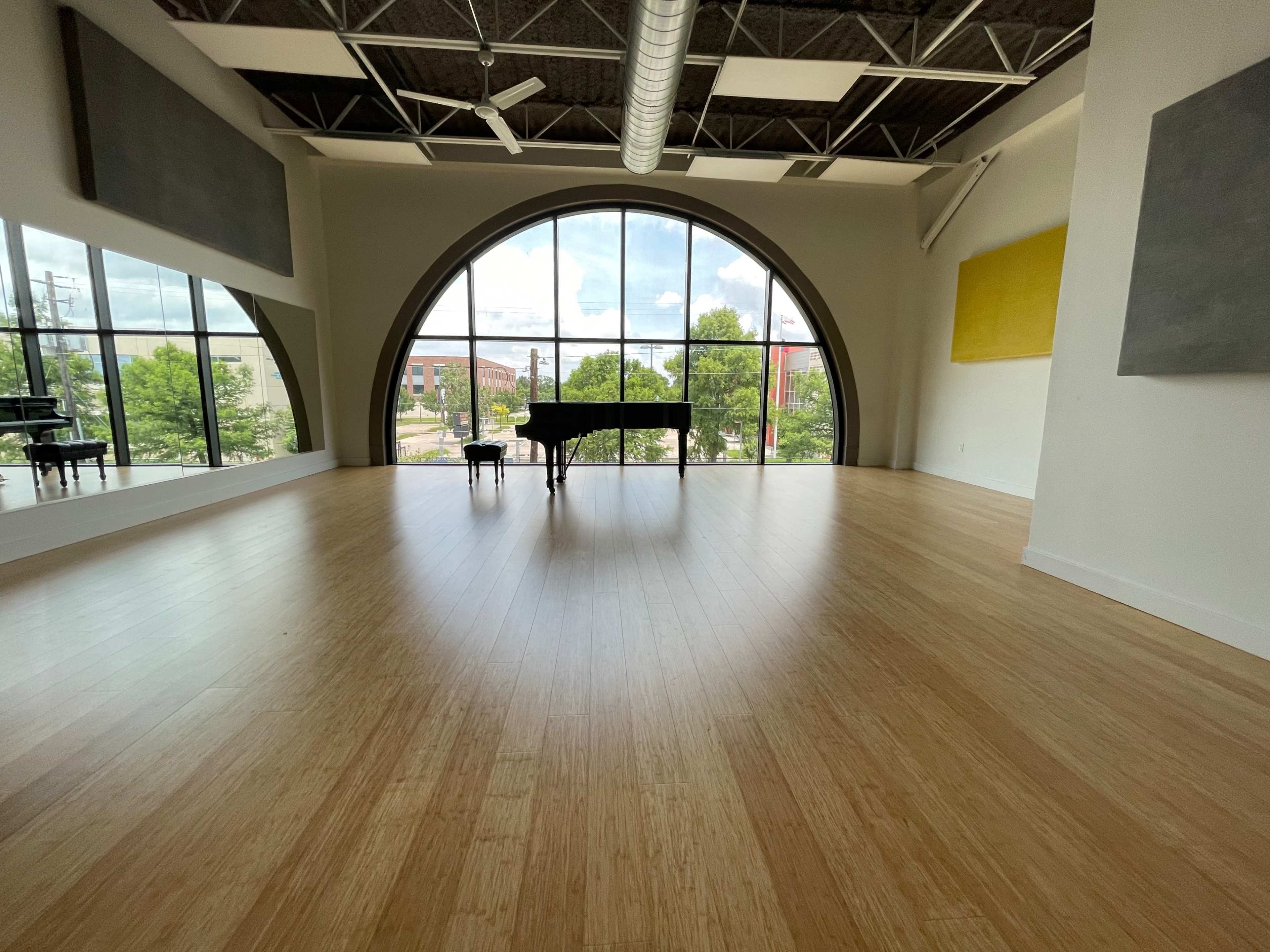 The image shows a spacious room with a large arched window, a grand piano, and wooden flooring.
