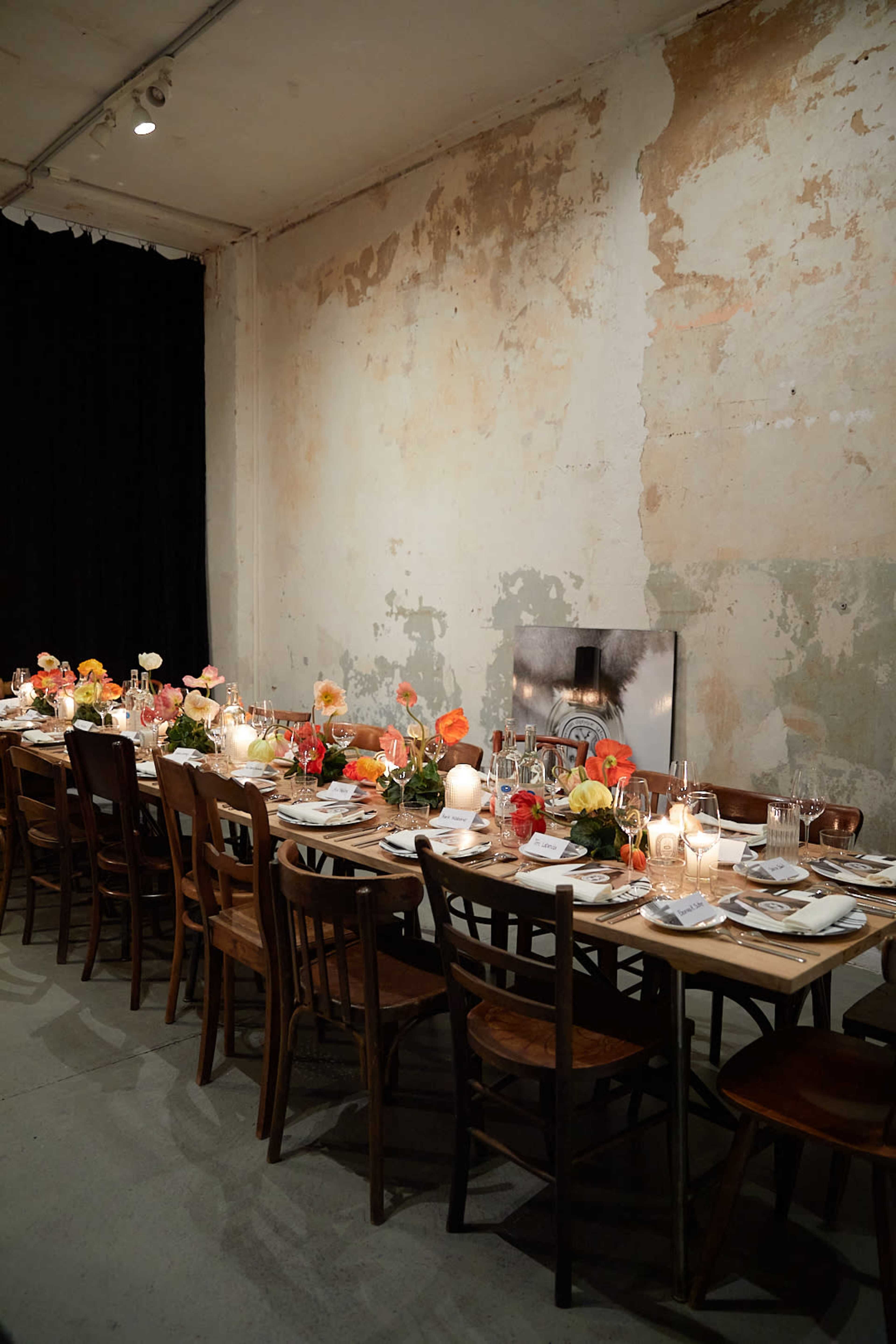 A long, elegantly set dinner table is adorned with colorful flowers and candles against a backdrop of a weathered, peeling wall.