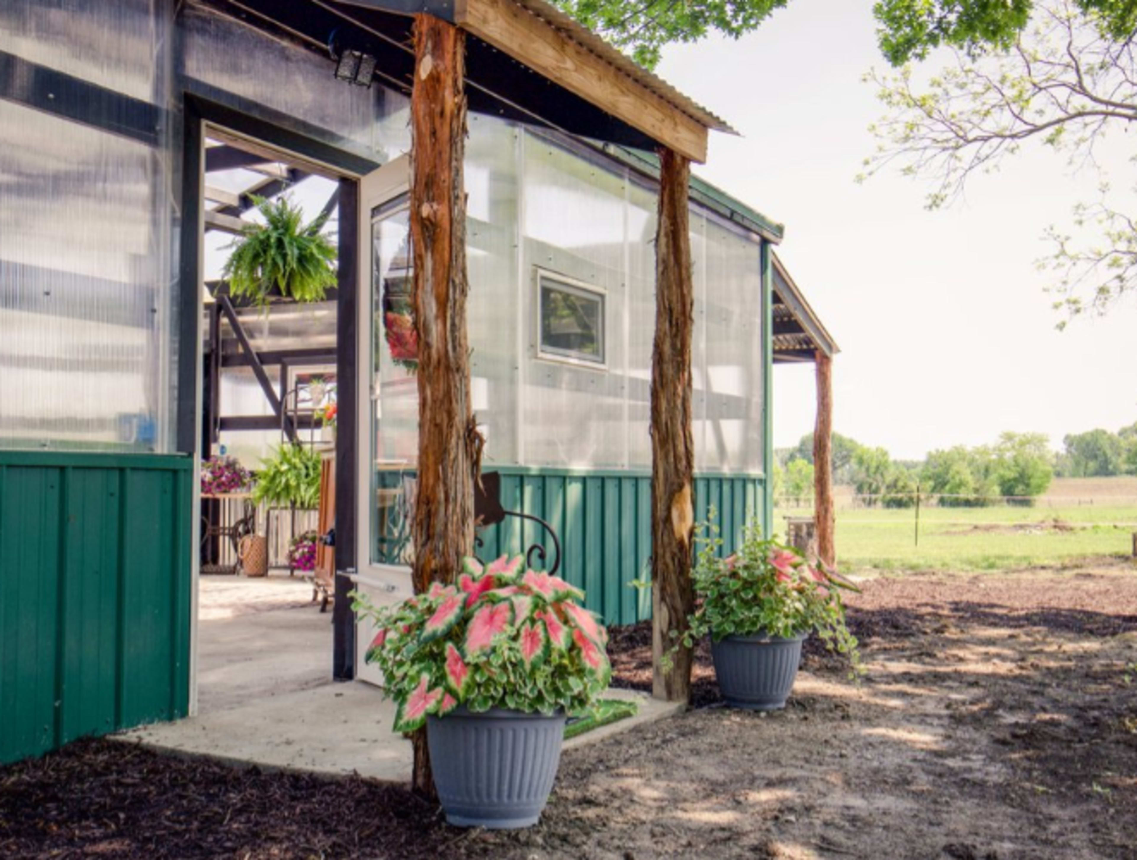A greenhouse with a clear plastic exterior features potted plants at its entrance and opens to a field in the background.