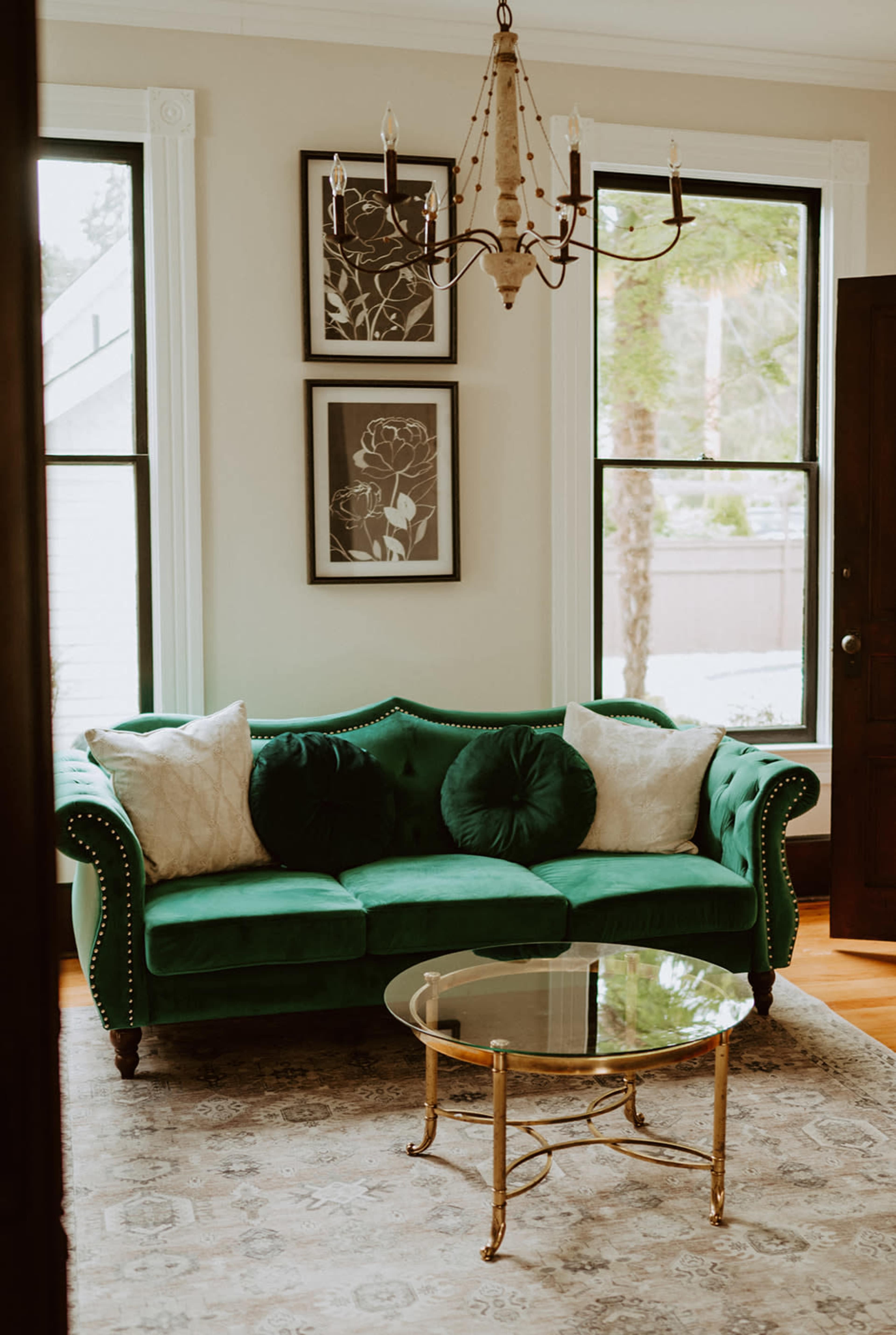 The image features a green velvet sofa with decorative pillows, a glass-top coffee table, and framed wall art in a well-lit room with large windows.