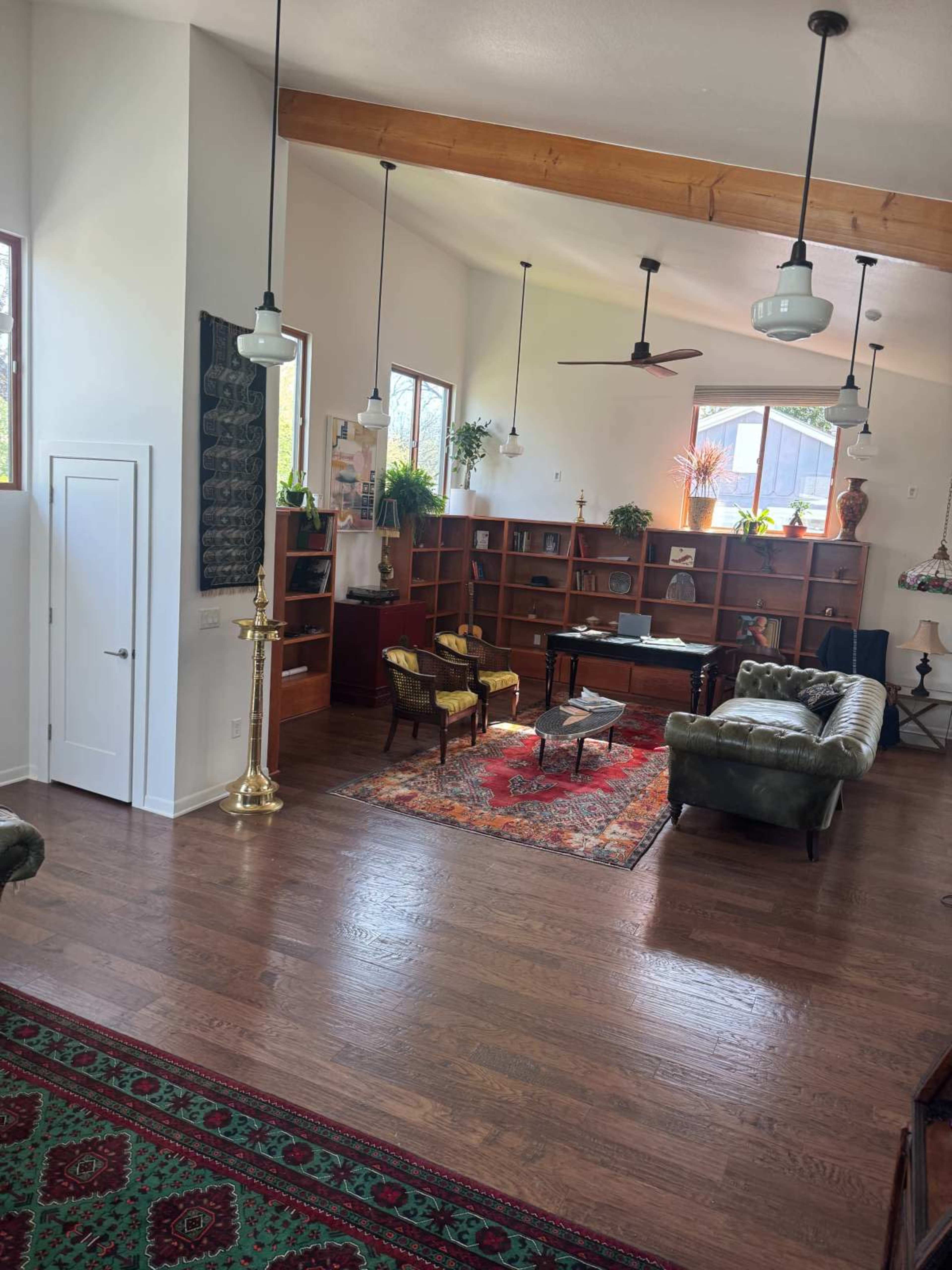The image shows a spacious living room featuring a mix of wooden furniture, a large bookshelf, plants, and a patterned rug on the hardwood floor.