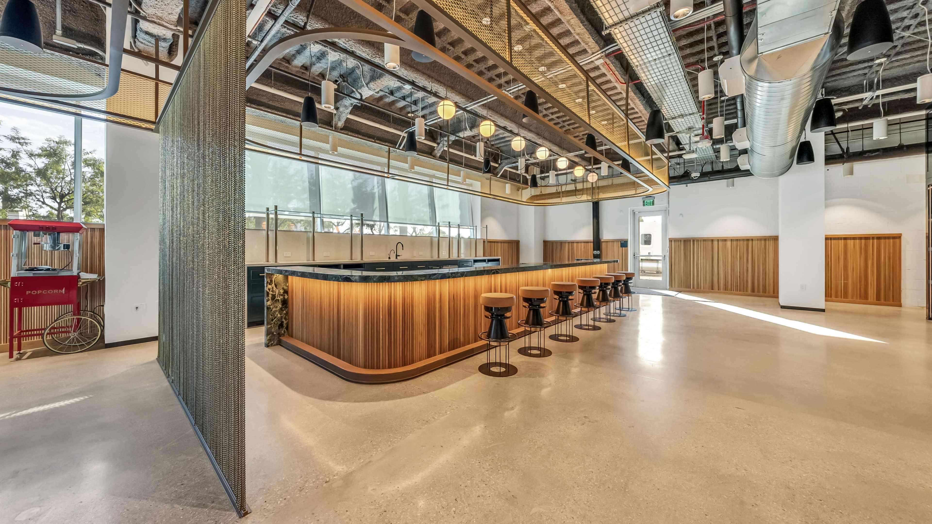 A modern interior space featuring a large bar with wooden accents, surrounded by stools and large windows allowing natural light.