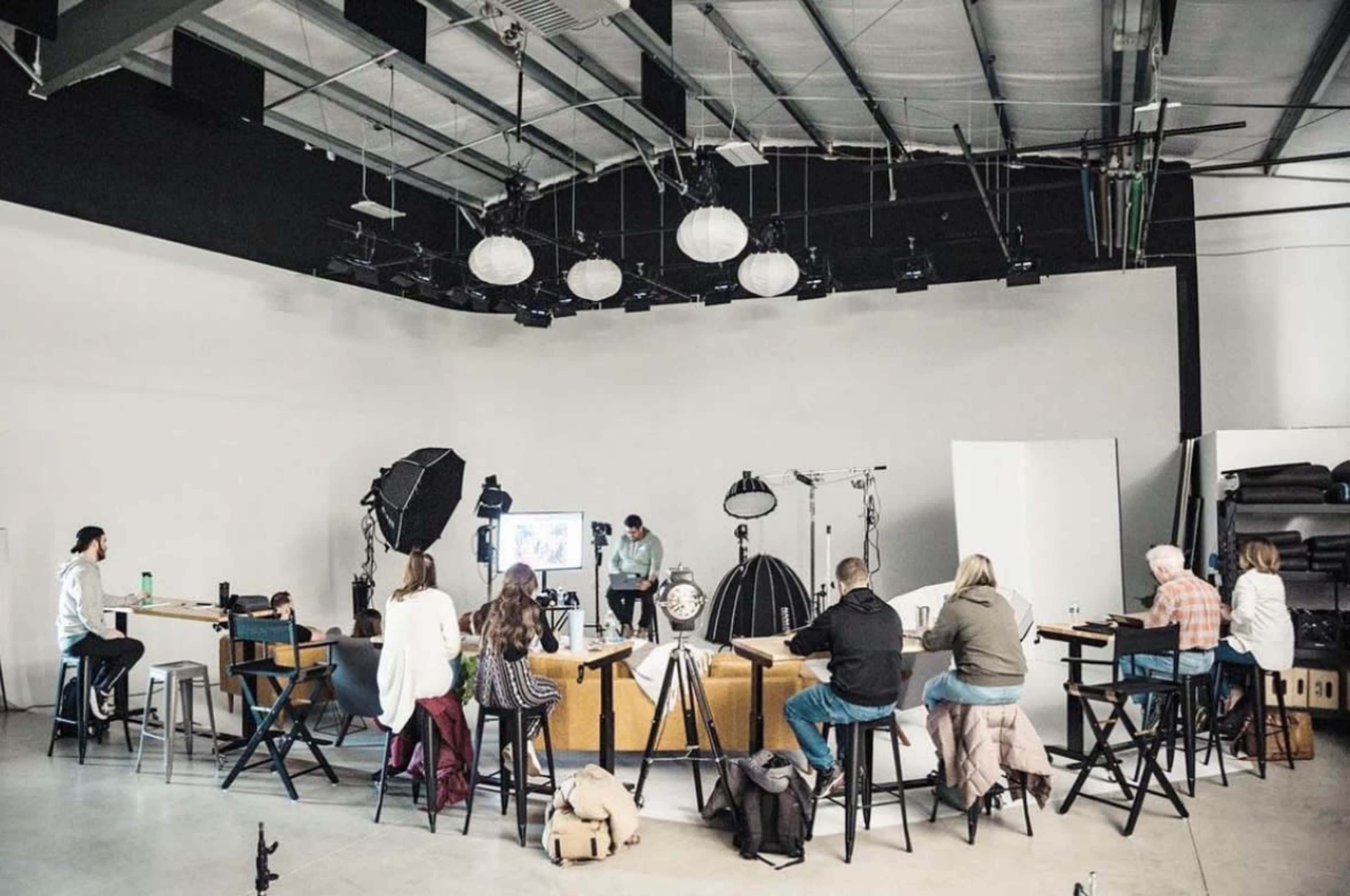 A group of people sits at a long table in a well-lit photography studio, observing a presentation on a screen while surrounded by various photography equipment.