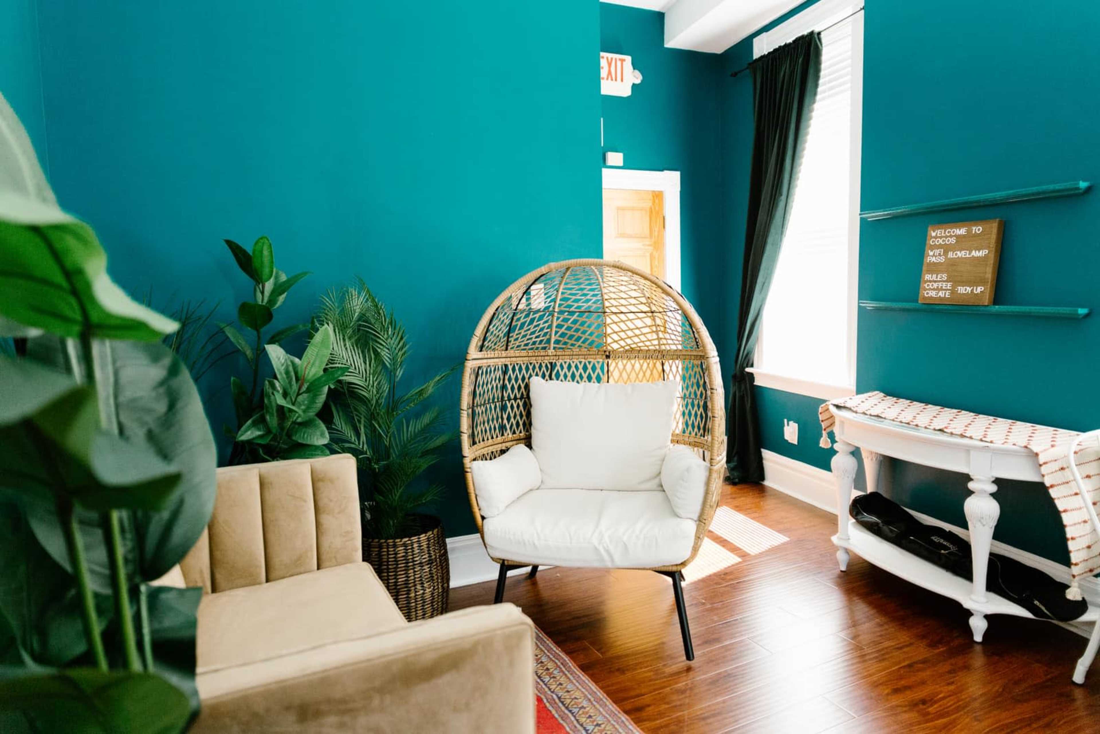 The image shows a cozy room with teal walls, featuring a rattan hanging chair, a beige sofa, and decorative plants.