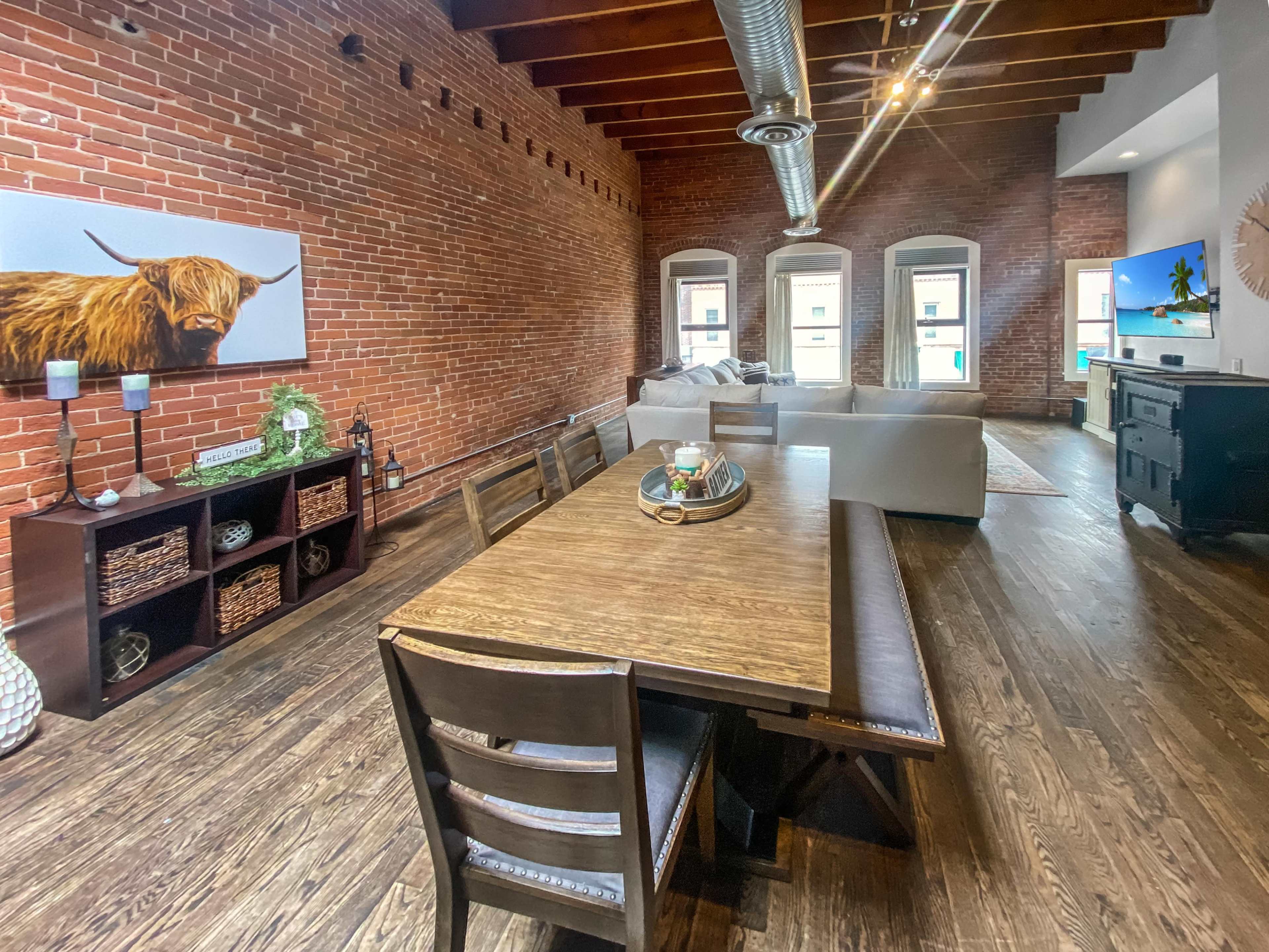 The image shows a spacious living and dining area featuring exposed brick walls, a wooden dining table with chairs, and a large sectional sofa near windows.