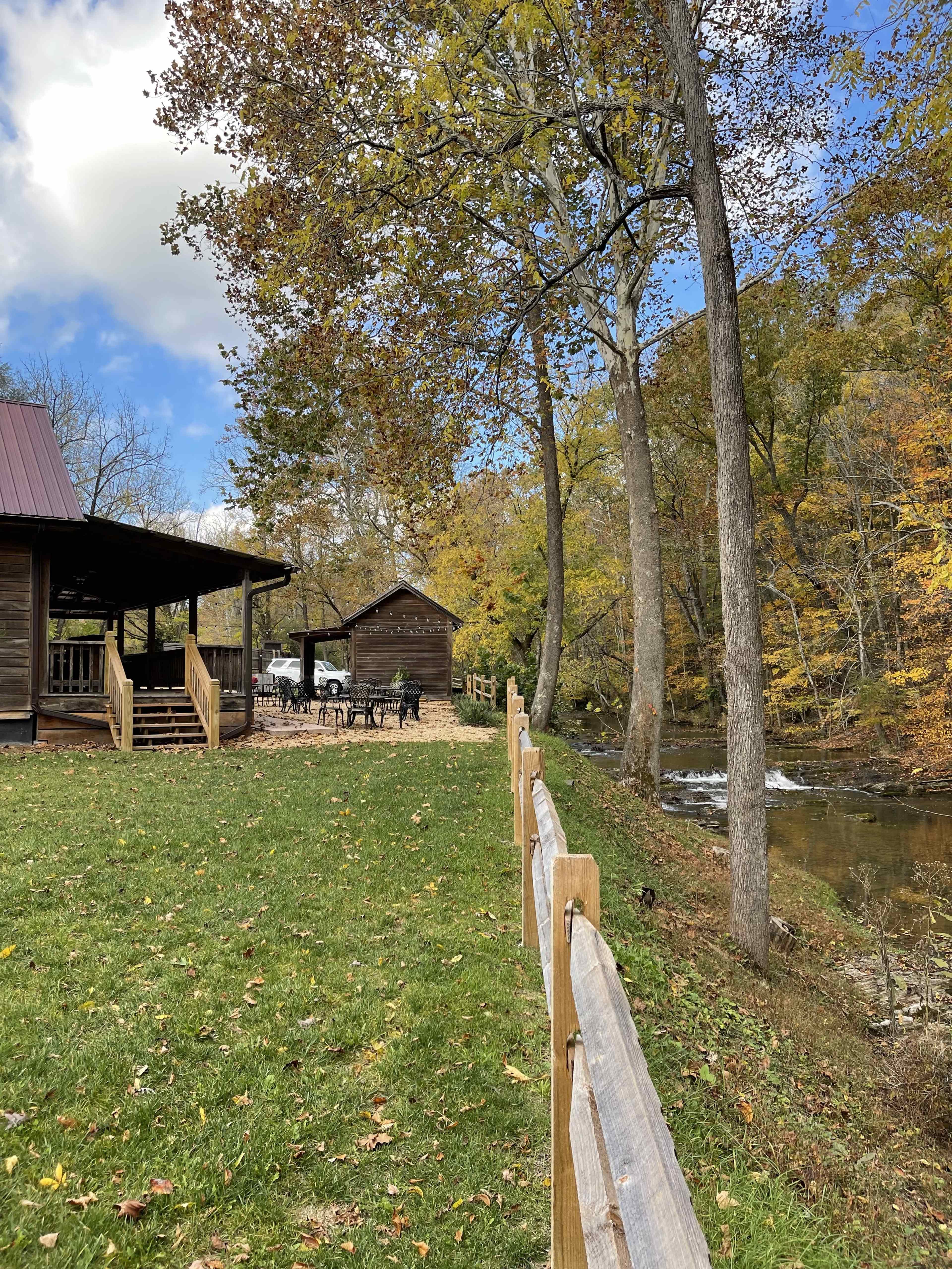 A rustic wooden cabin sits beside a gently flowing stream, with a grassy area and a wooden fence in the foreground.