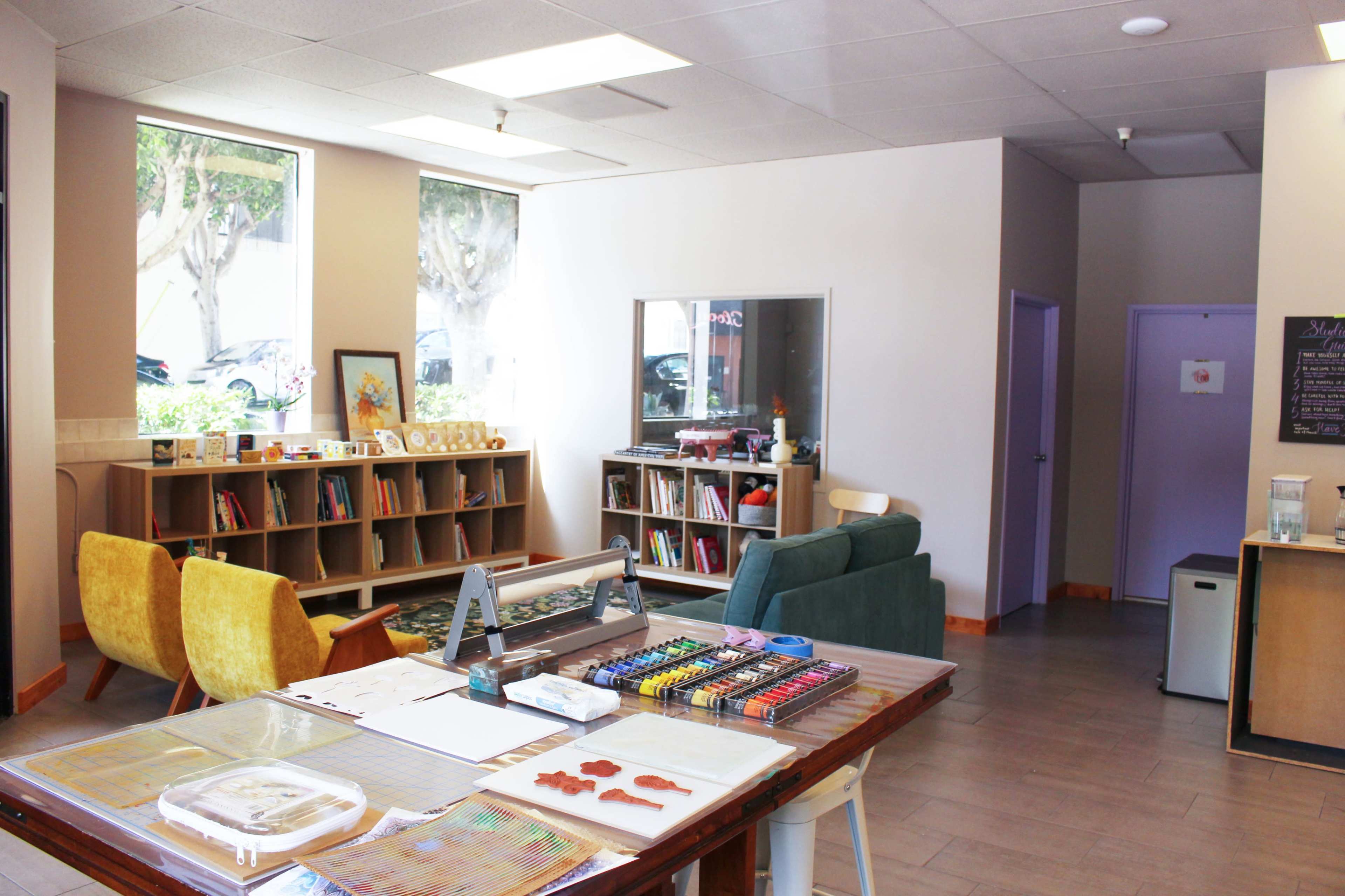 The image shows a bright, open space with a table covered in art supplies, a bookshelf filled with books, and seating areas featuring a green sofa and yellow chairs.