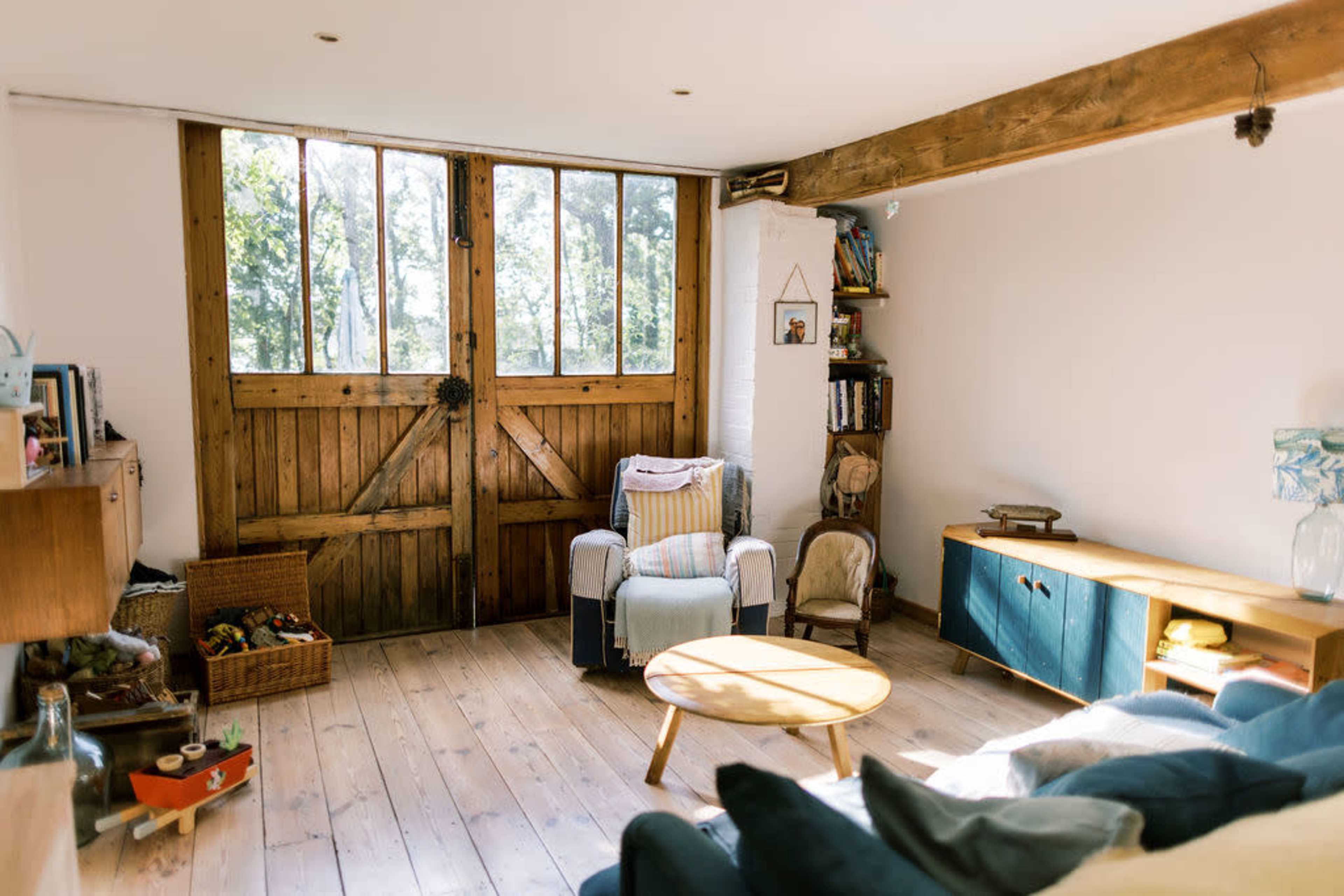 A cozy living room features a wooden door with large windows, a light wooden floor, and a mix of seating and shelving units.