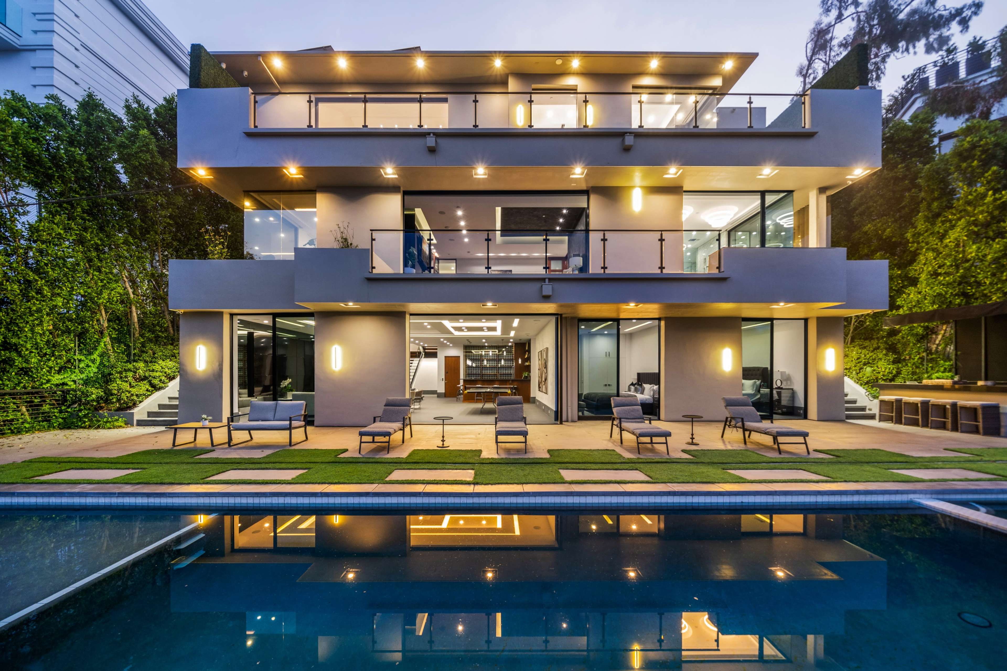 A modern three-story house with large glass windows, surrounded by greenery and featuring a swimming pool in the foreground.