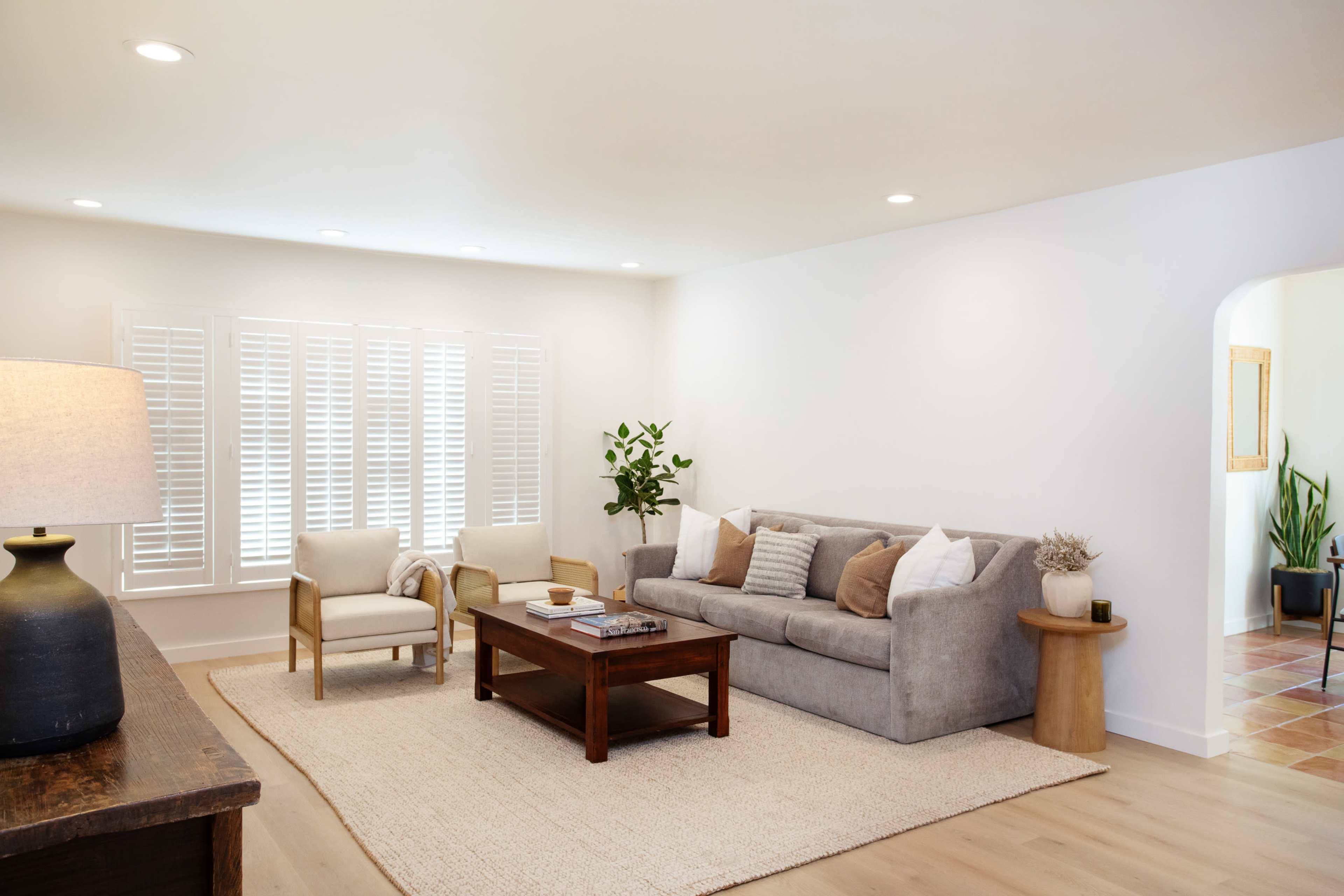 The living room features a gray sofa, two armchairs, a coffee table, and a light-colored rug with window shutters in the background.