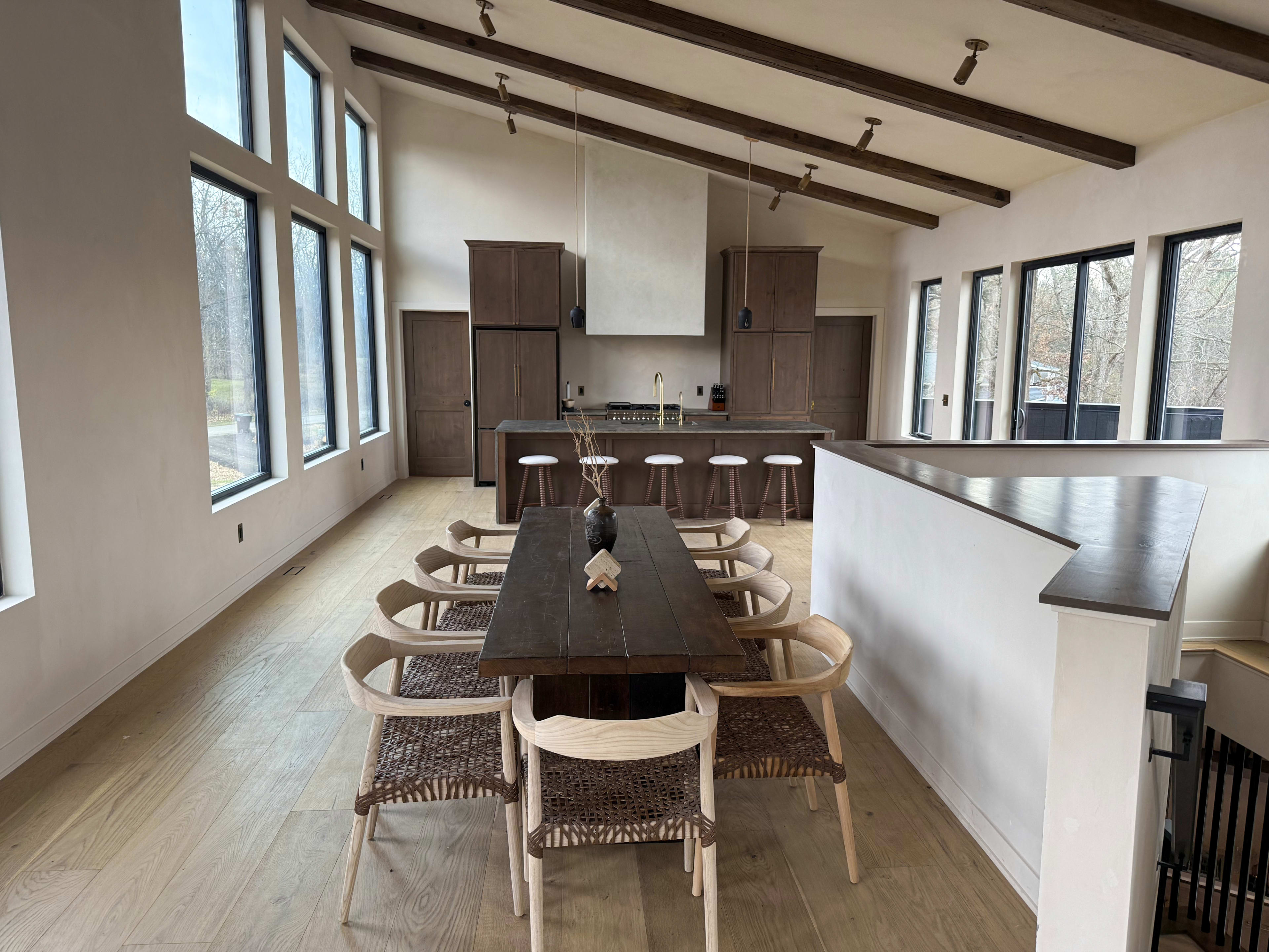 A long wooden table with woven chairs sits in a spacious dining area adjacent to a modern kitchen with large windows.