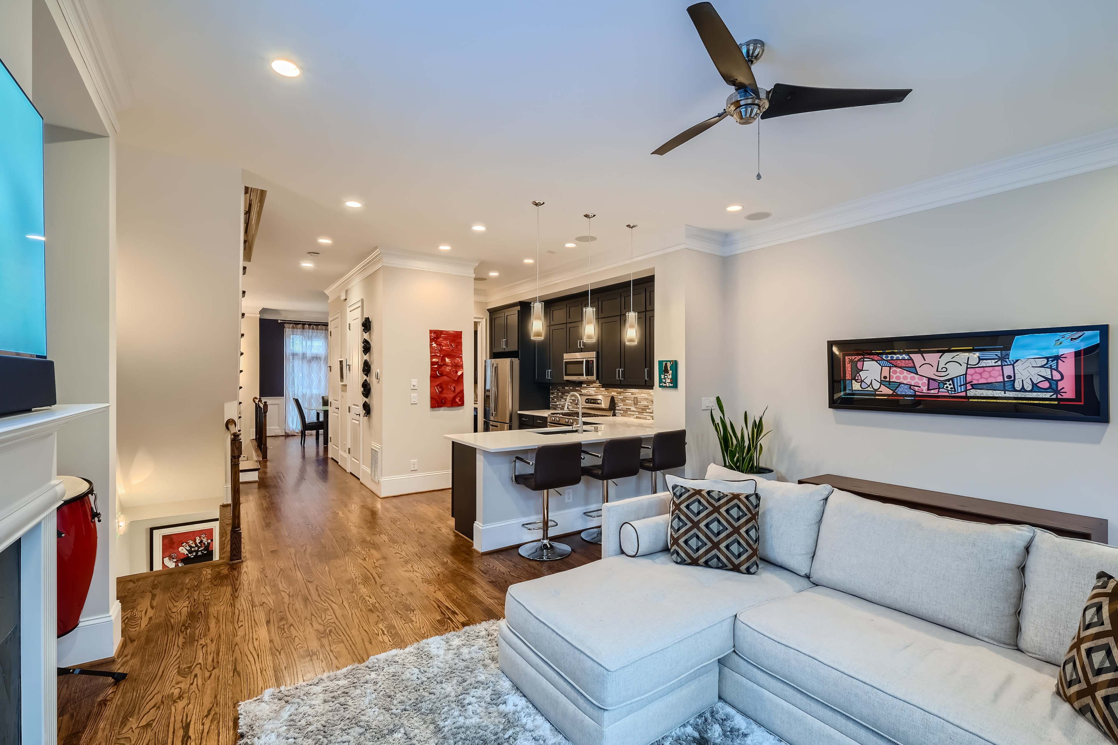 The image shows a modern living room with a sectional couch, a kitchen area featuring a breakfast bar, and hardwood floors leading to a hallway.
