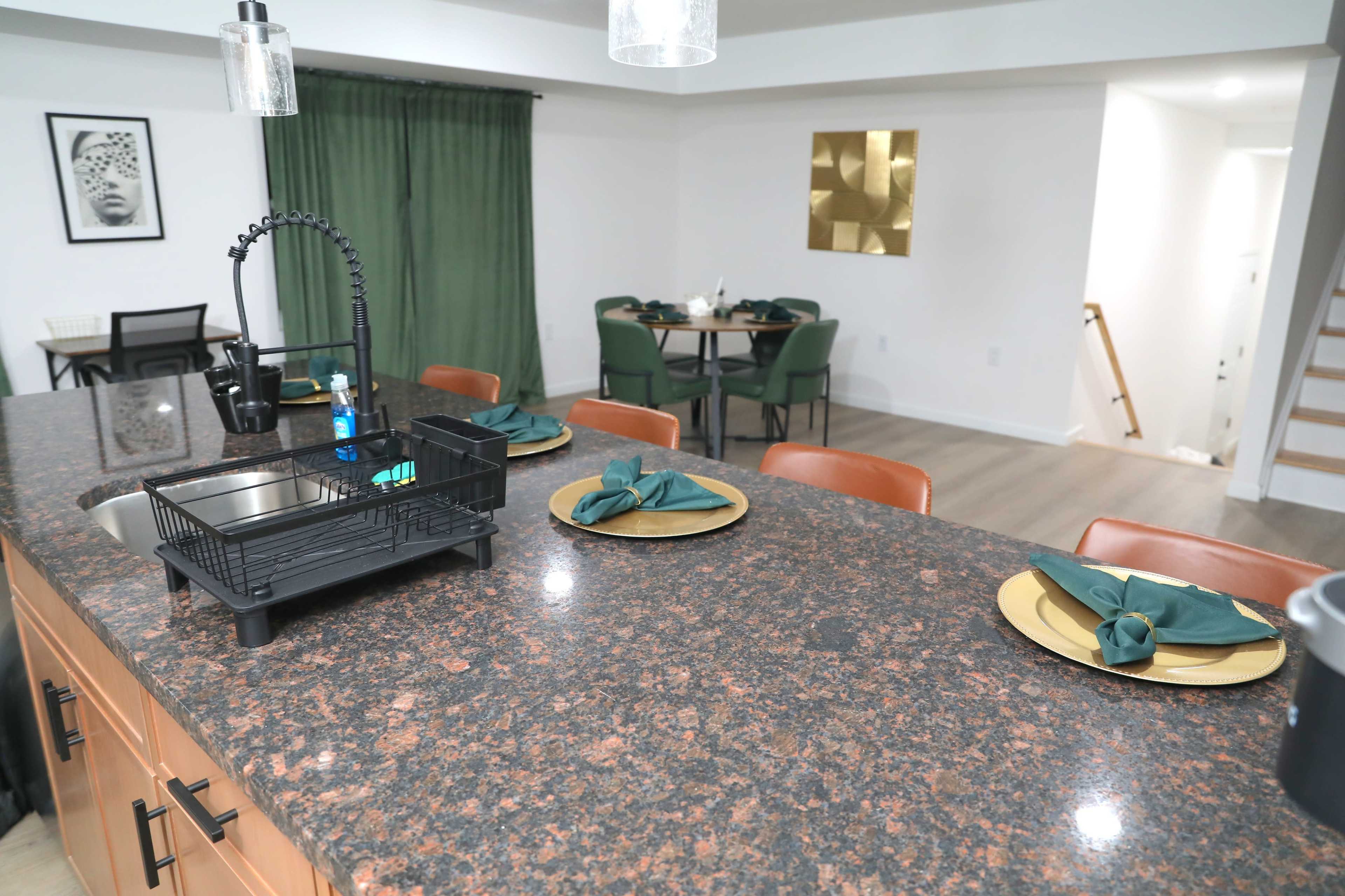The image shows a modern kitchen with a granite countertop, featuring a drying rack and neatly arranged dinner plates, while a dining area with green curtains and a gold wall decoration can be seen in the background.