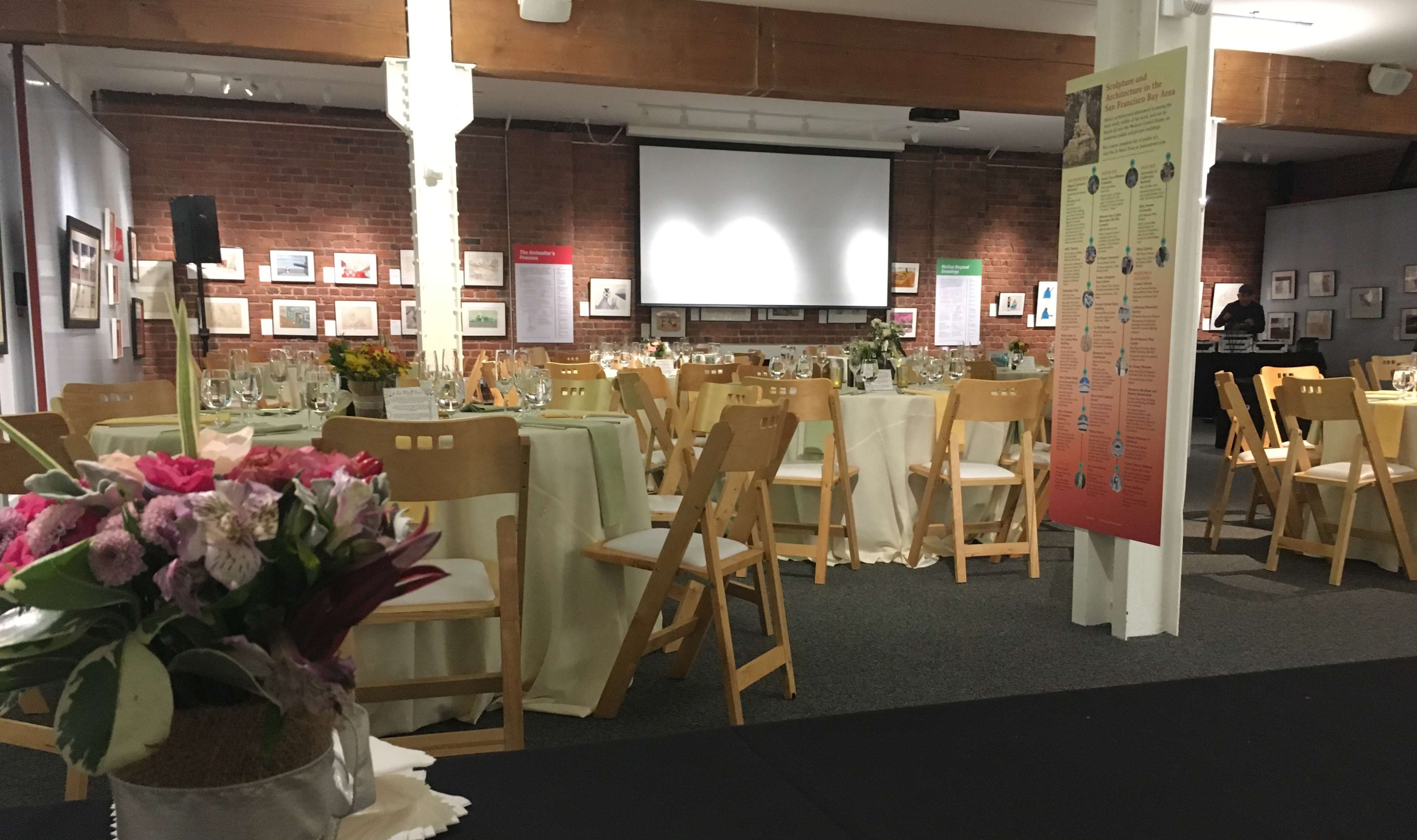 A dining setup is arranged in a hall with tables dressed in light-colored linens, featuring floral centerpieces and a projector screen in the background.