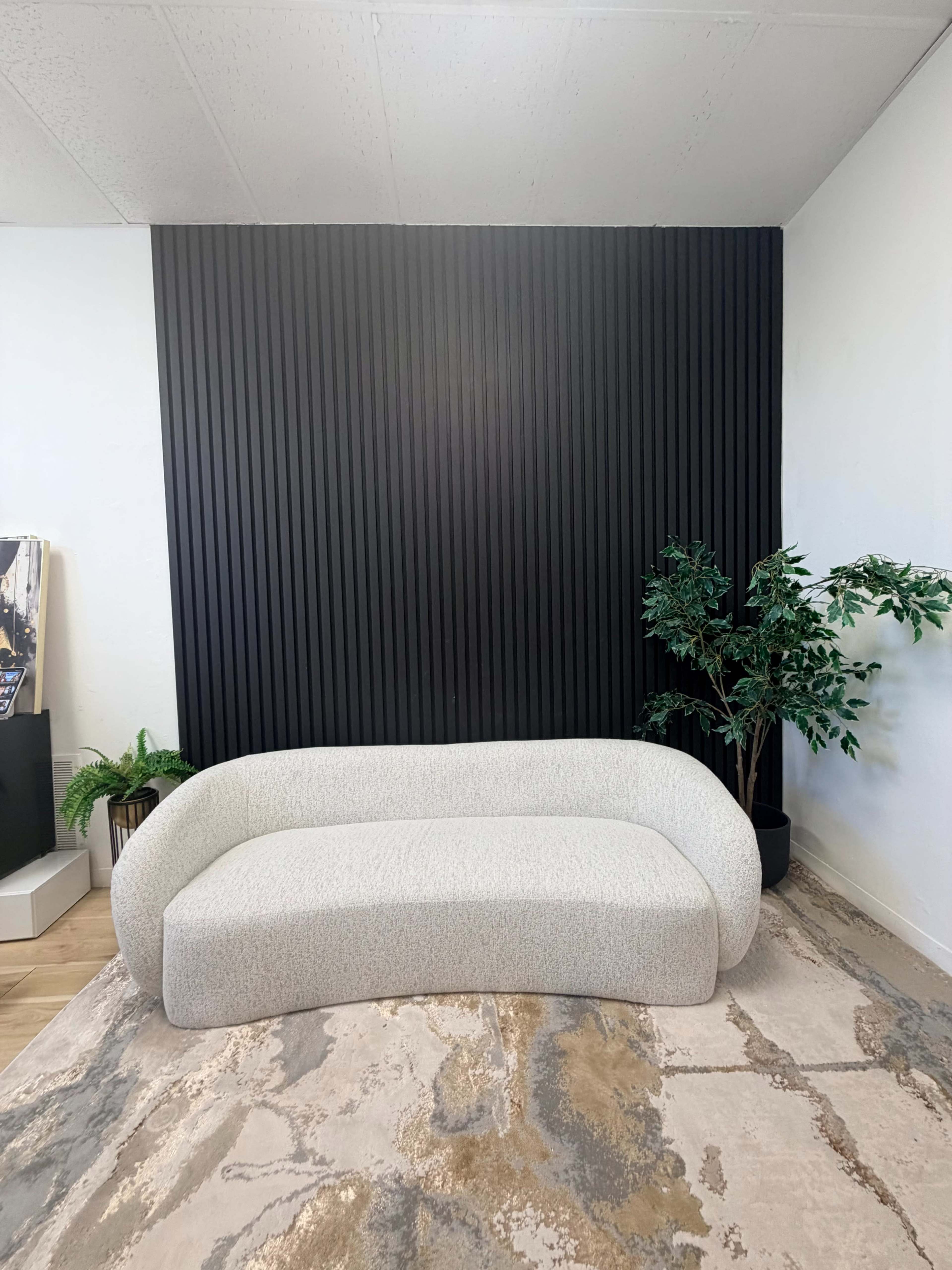 The image shows a light-colored, curved sofa positioned in front of a black, vertically slatted wall, with a potted plant to the right and a textured, neutral-toned rug on the floor.