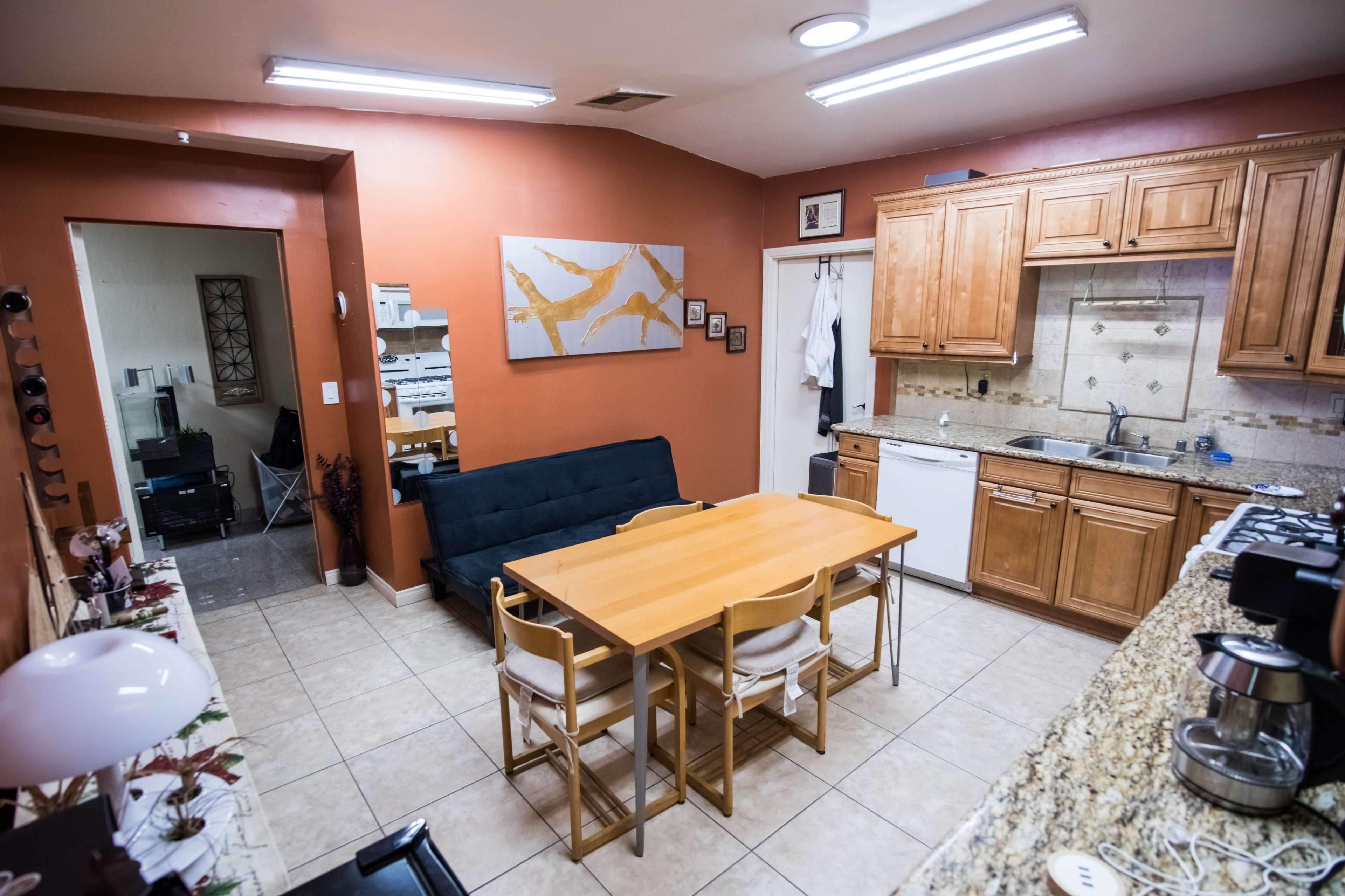 A kitchen and dining area features wooden cabinetry, a granite countertop, a dining table with white chairs, and a black sofa against an orange wall.