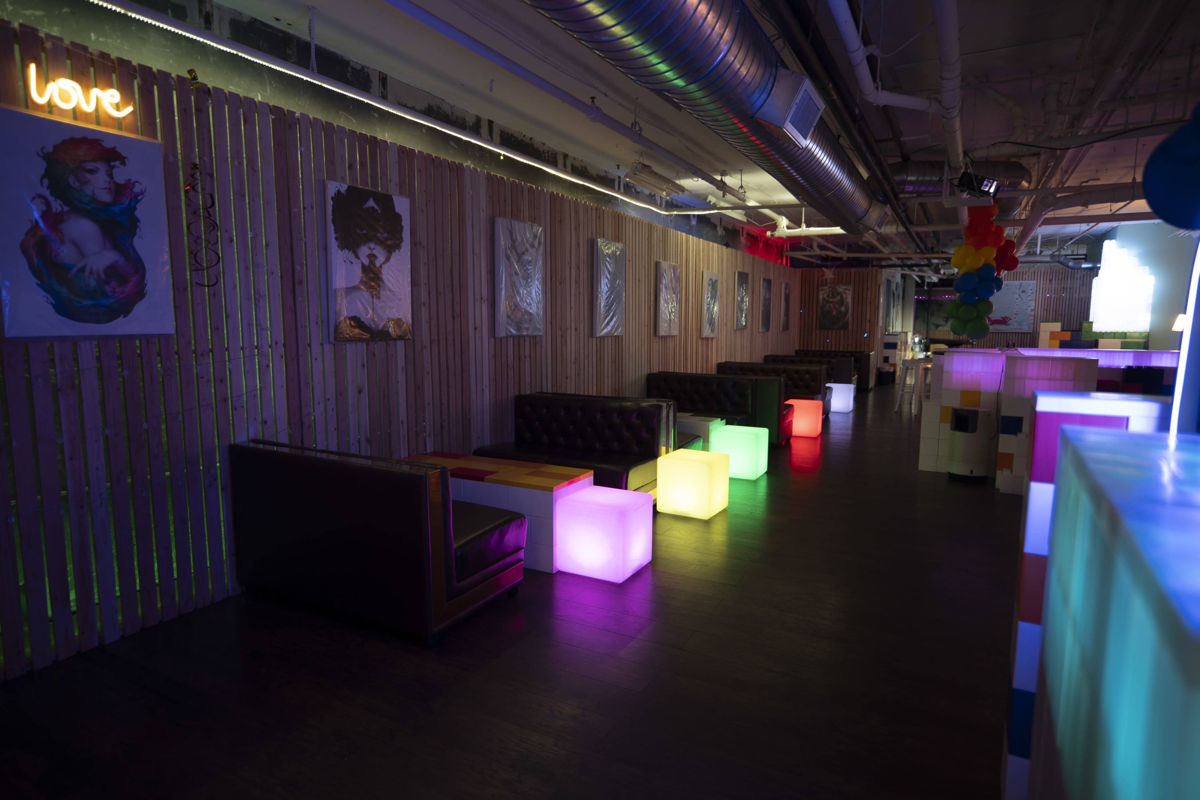 A dimly lit lounge area with seating and colorful illuminated tables along the walls decorated with art.