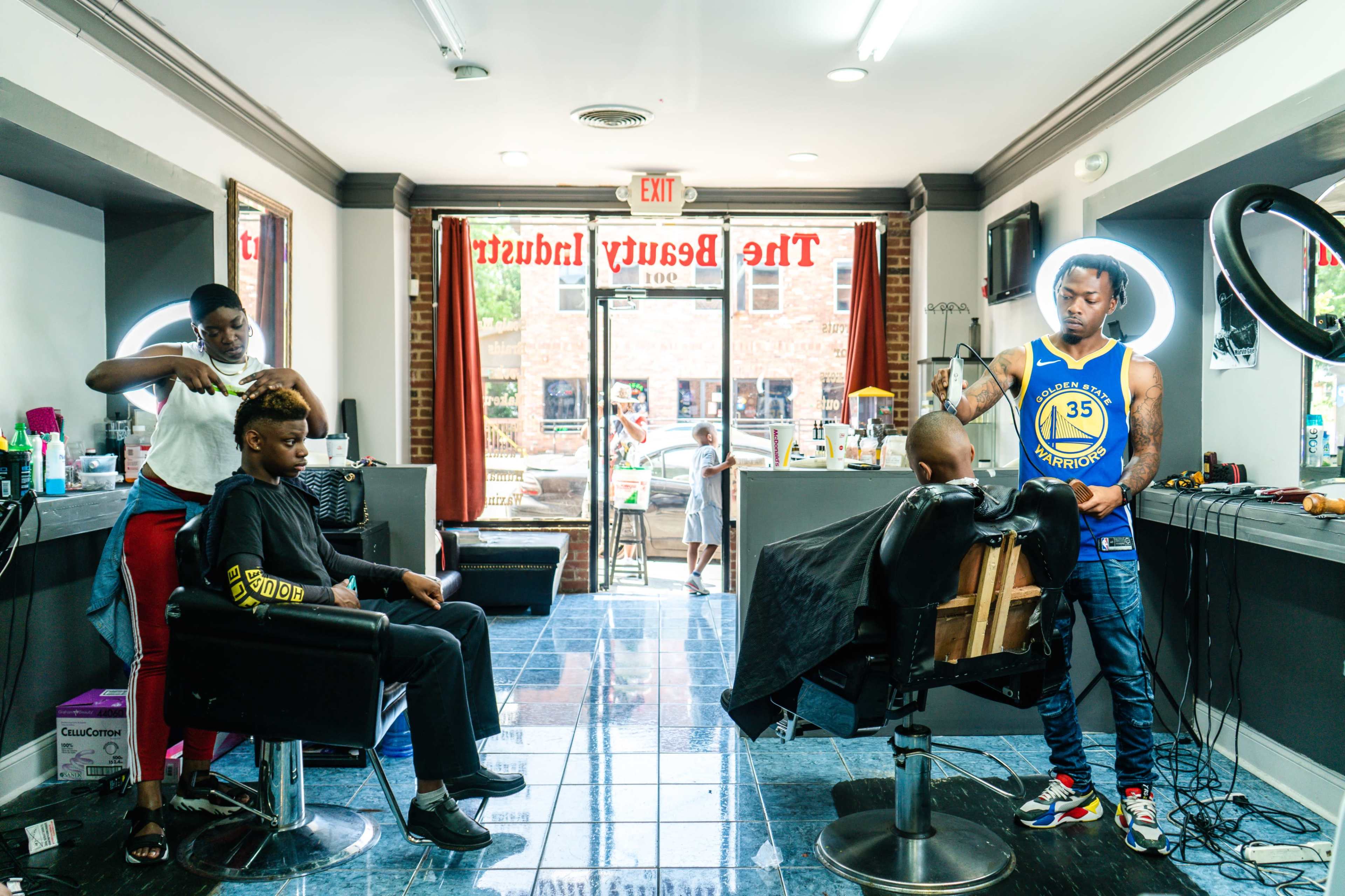 A barber shop interior features two clients in chairs, with one barber cutting hair and another stylist attending to a different client.
