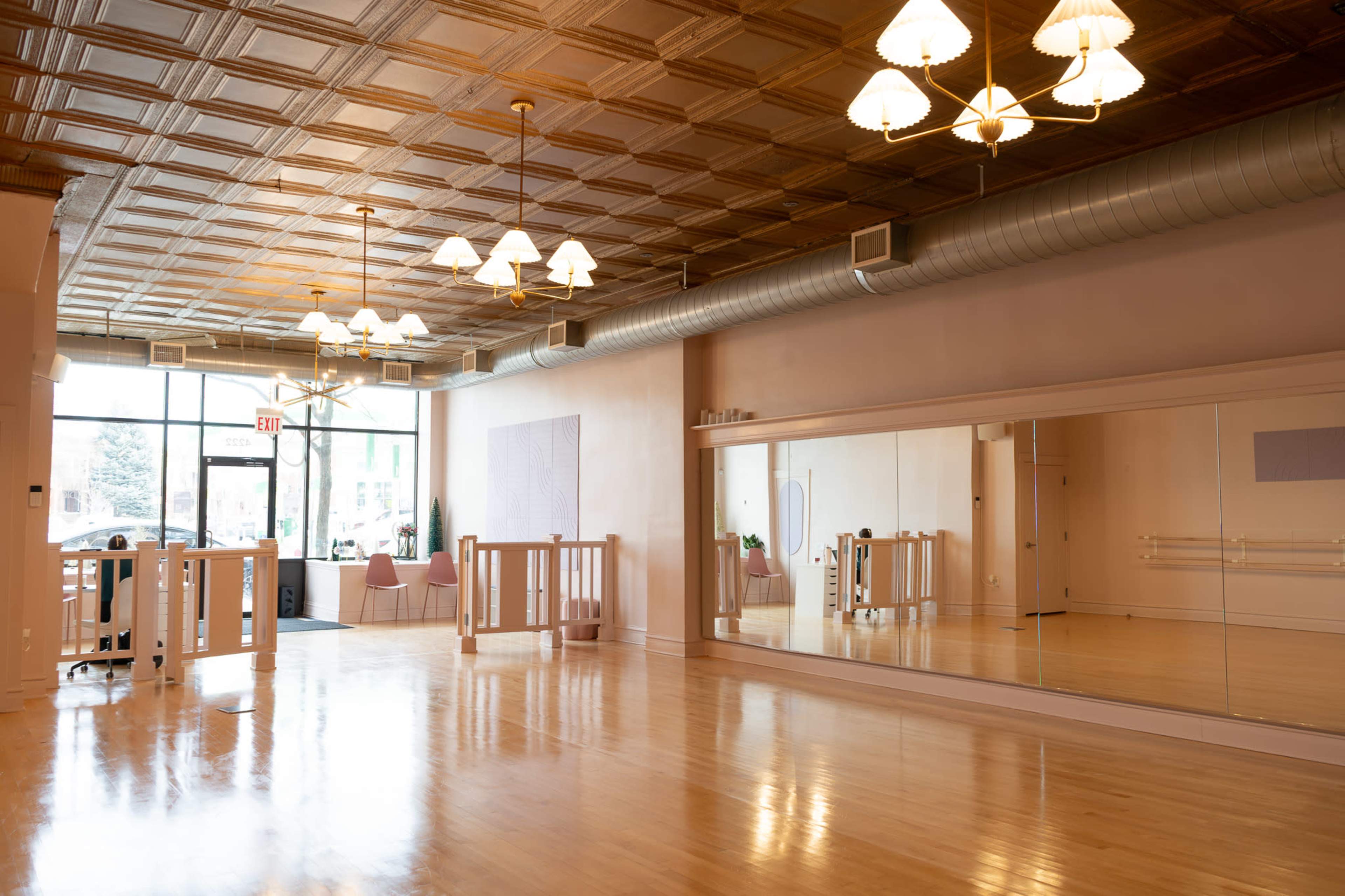 The image shows a spacious, well-lit dance studio with a hardwood floor, large mirrors along one wall, and decorative lighting fixtures on a textured ceiling.