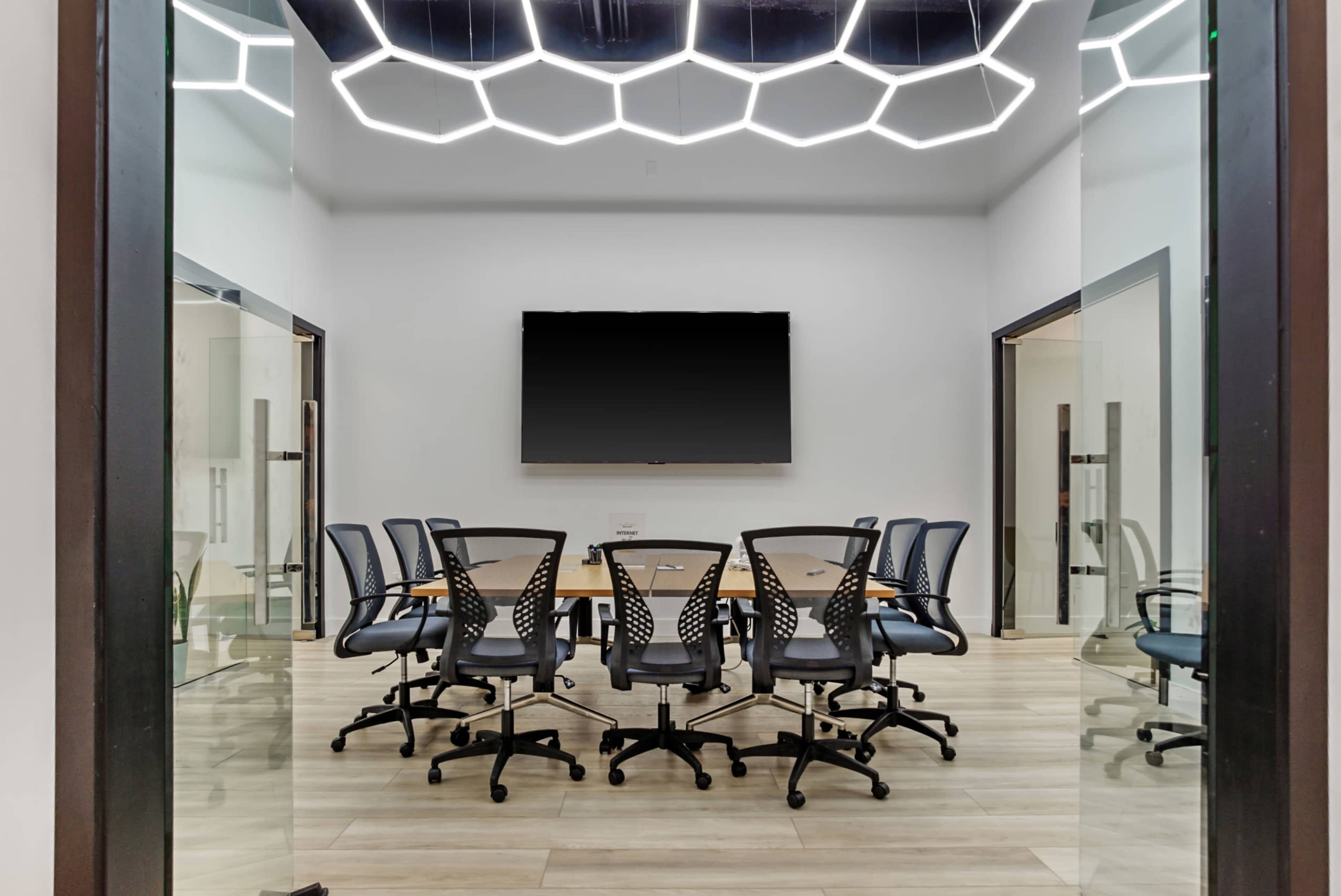 A modern conference room features a large table surrounded by ergonomic chairs, with a wall-mounted screen and hexagonal light fixtures overhead.