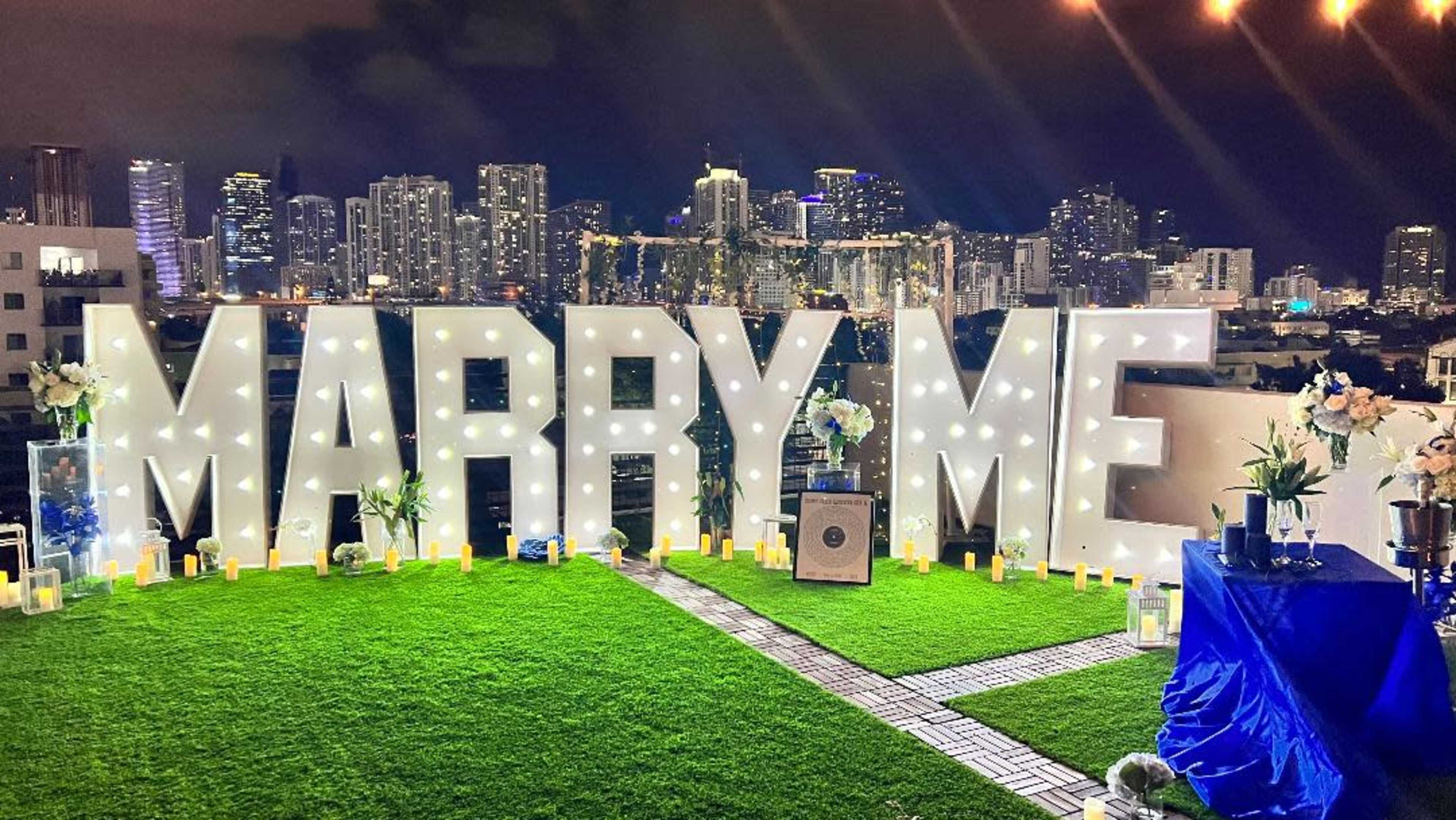 The scene features a large "MARRY ME" display illuminated with lights on a rooftop, surrounded by candles and flowers, with a city skyline in the background.