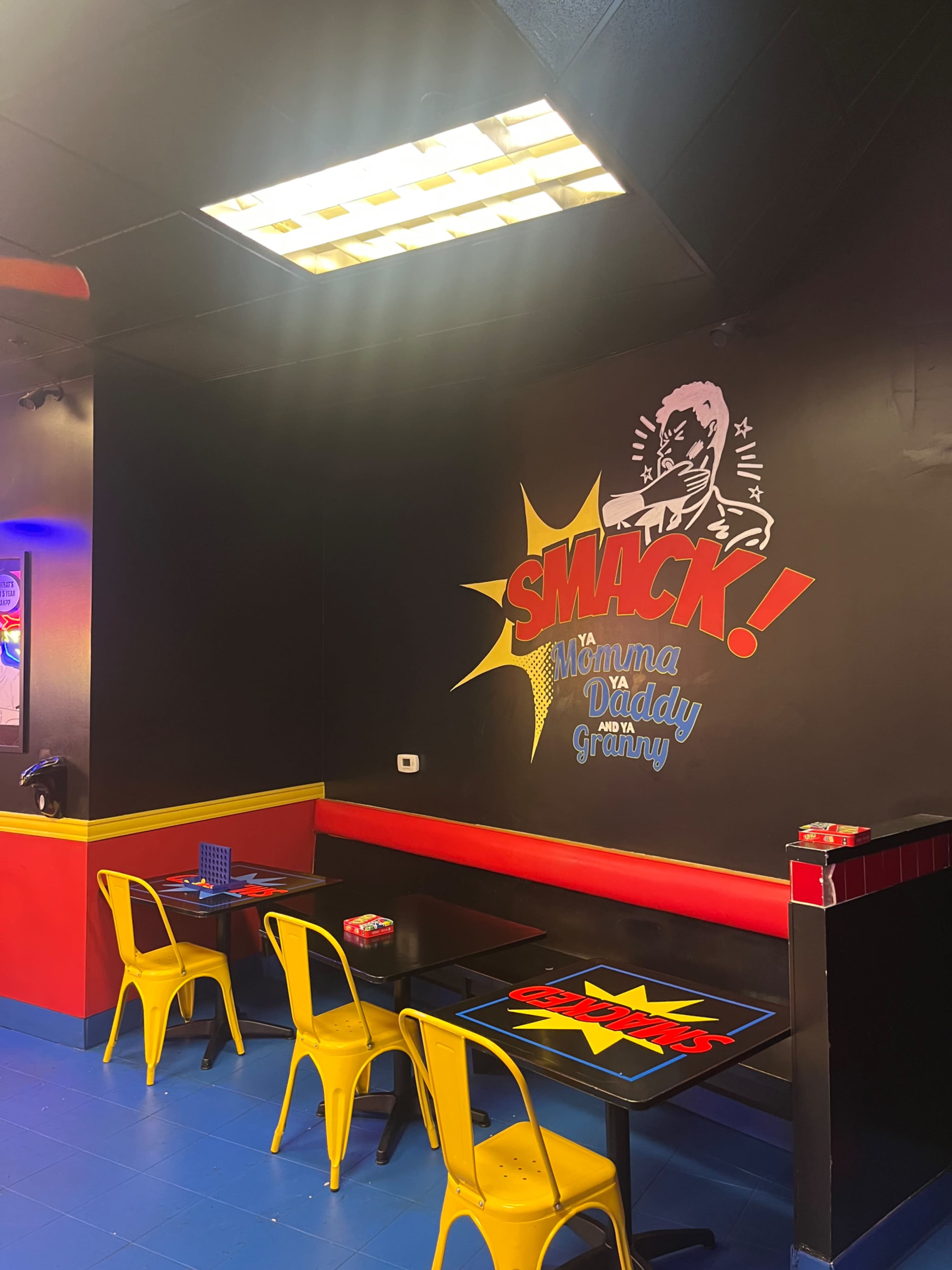The image shows a colorful fast-food restaurant interior featuring yellow chairs and tables with a comic-style "SMACK!" graphic on the wall.