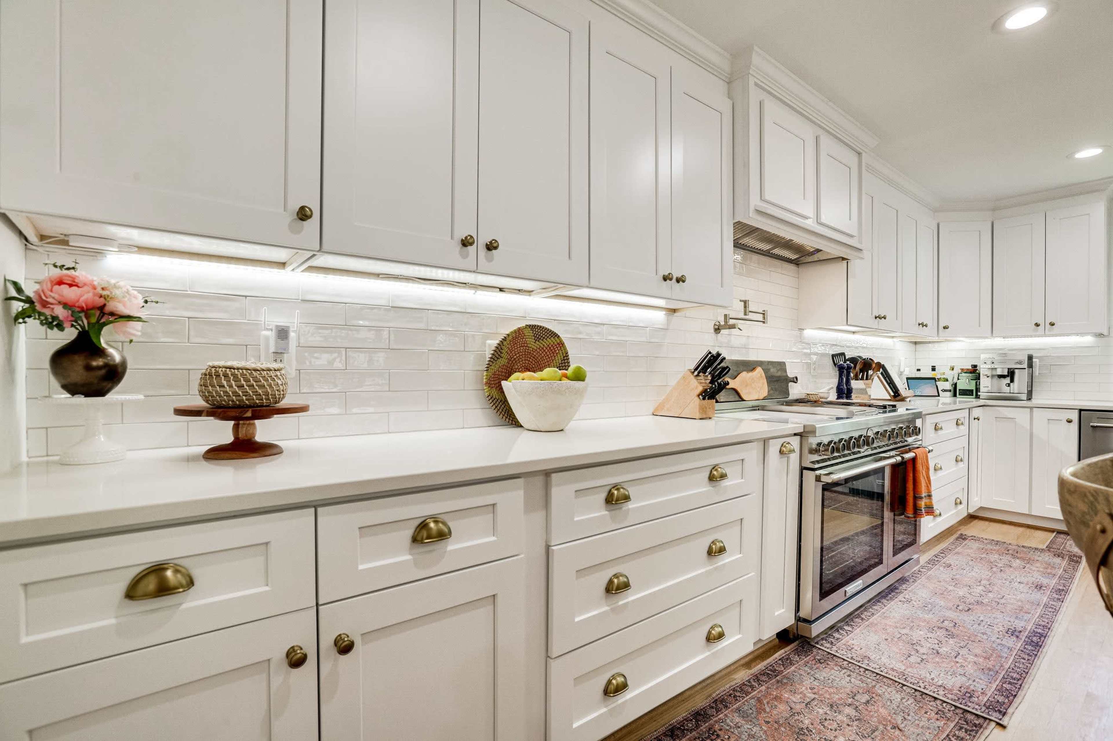 The image shows a modern kitchen with white cabinets, a stainless steel range, and a decorative rug on the floor.