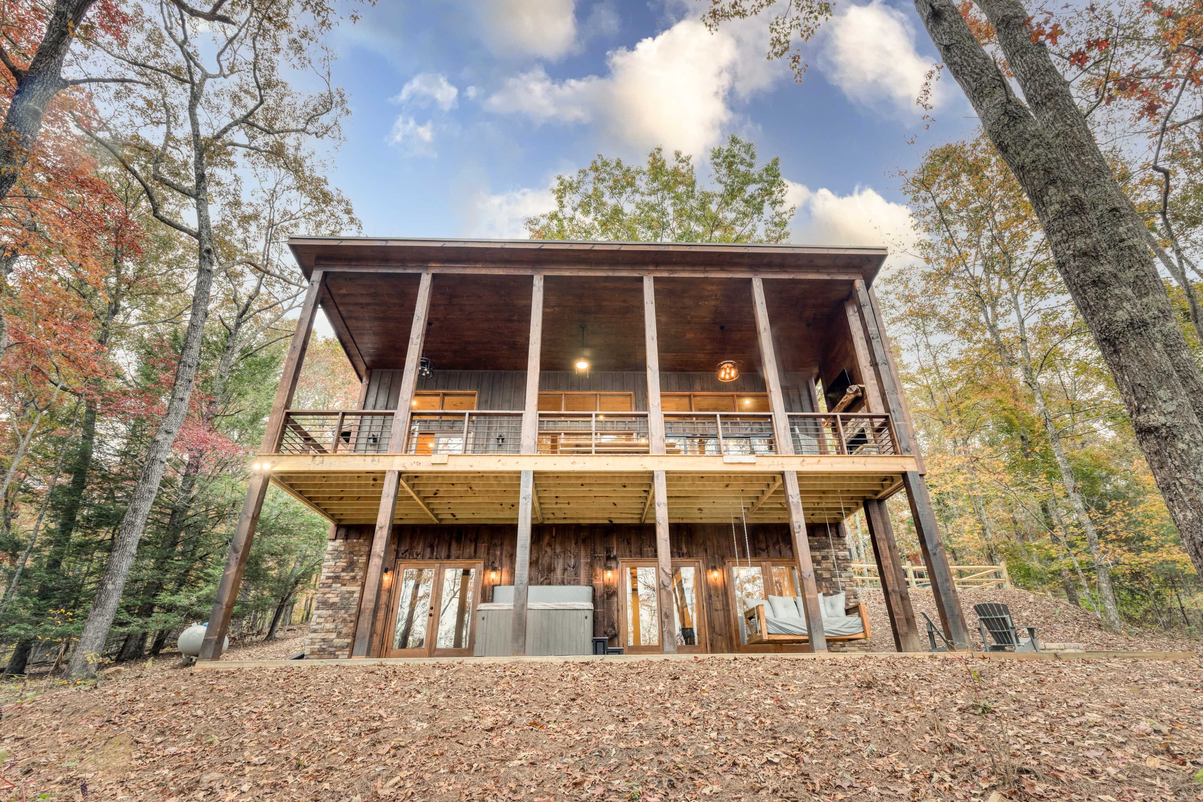 A two-story wooden cabin with a spacious deck overlooks a landscaped area surrounded by trees and fallen leaves.