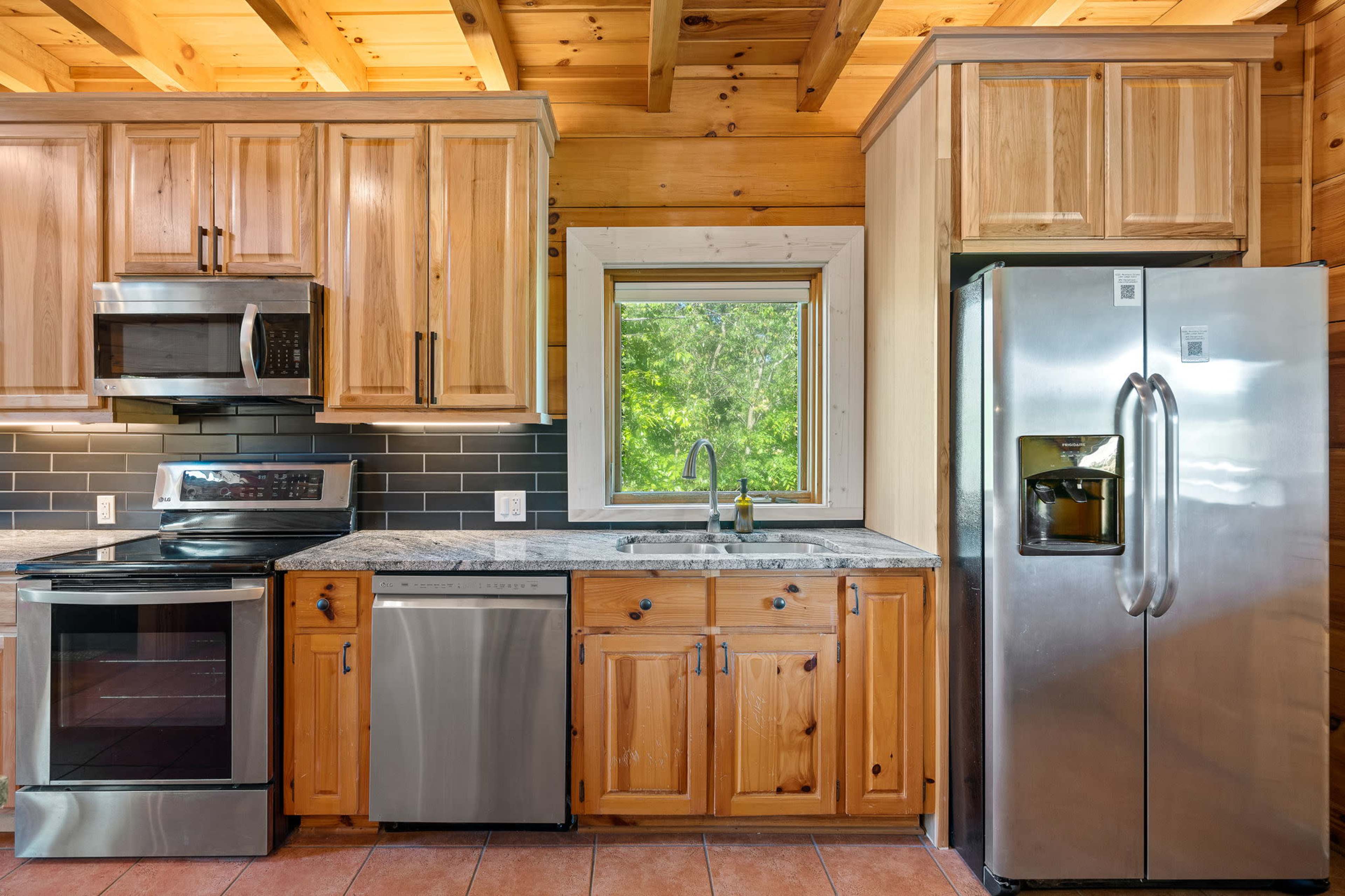 A modern kitchen features wooden cabinets, stainless steel appliances, and a window overlooking greenery.