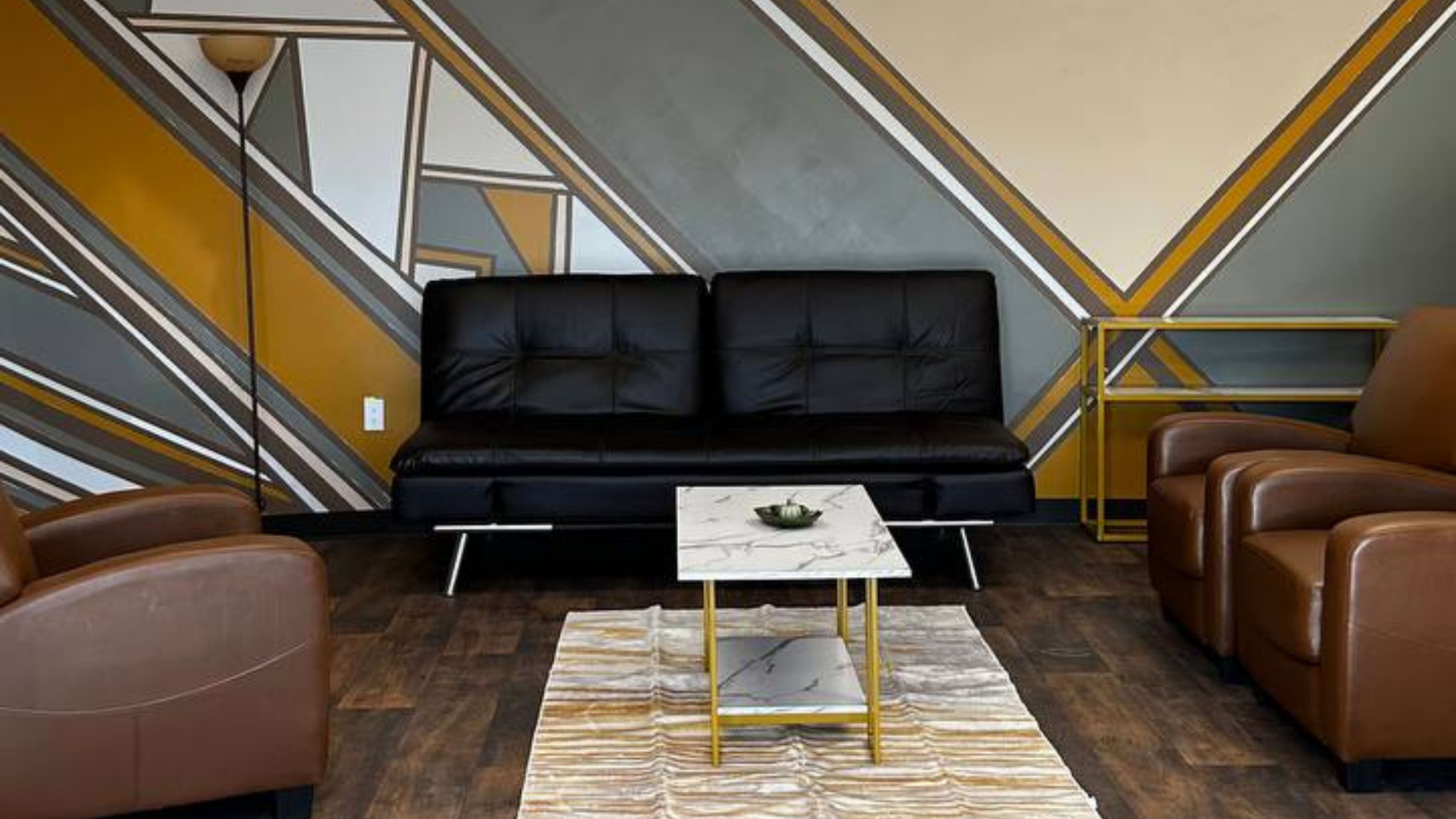 The image shows a modern seating area with a black sofa, brown leather chairs, a marble-topped coffee table, and geometric wall designs in gray and gold.